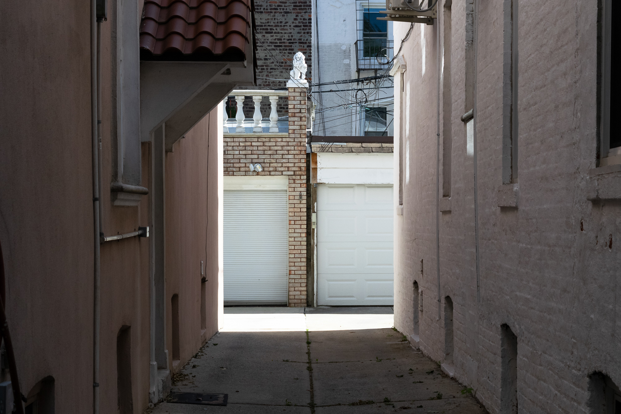 A shaded alley between two stucco and brick row houses opens onto a sunlit rear yard with two white garage doors, one roll-down and one paneled. A brick pier between them is capped with a small white lion statue beside a balustrade, with utility wires crossing the sky above.