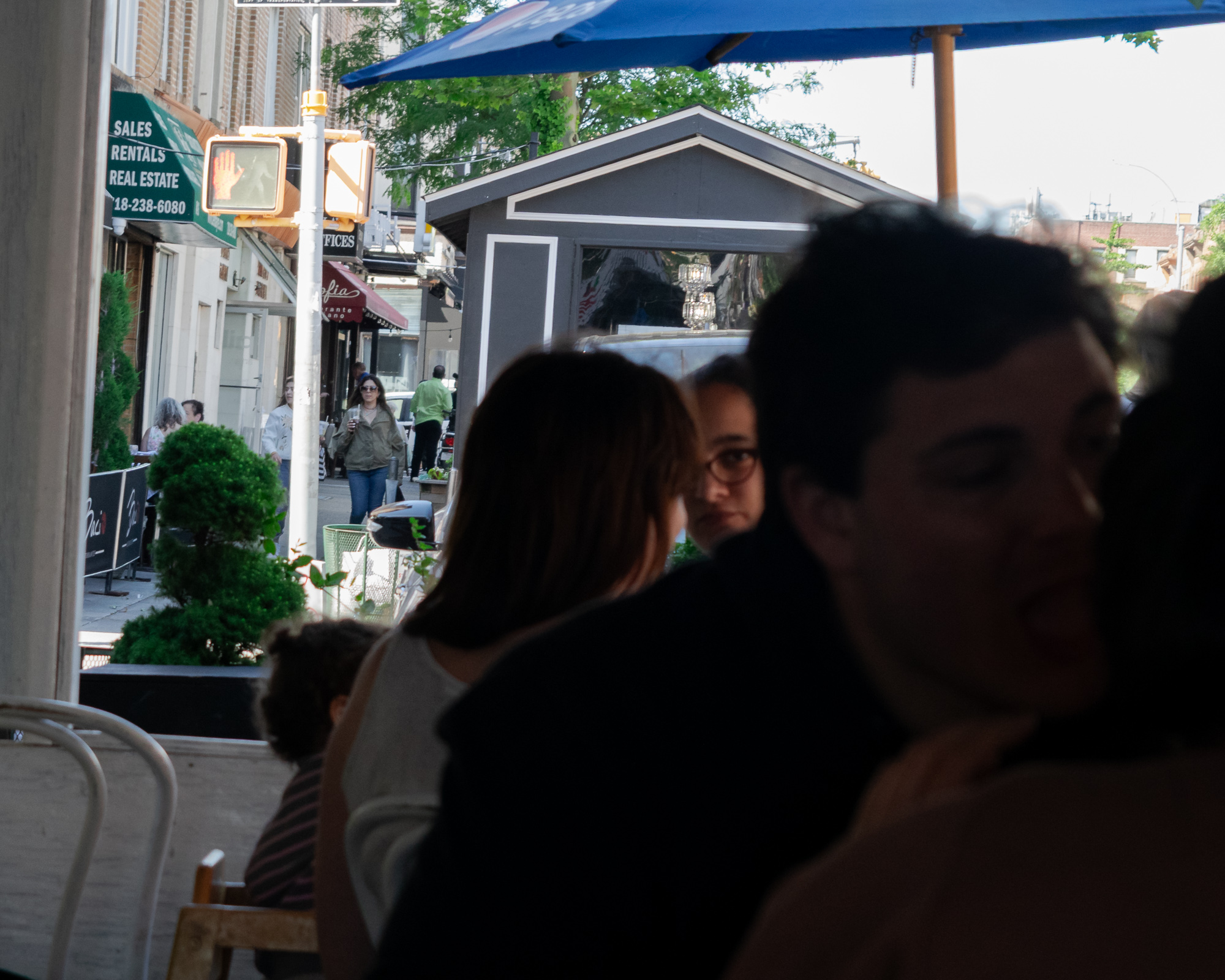 Shot from a shaded cafe interior looking out to a sunlit Brooklyn sidewalk, silhouetted heads and shoulders of seated diners fill the right and center of the frame while a woman in glasses turns and looks directly at the camera. Beyond them, a blue patio umbrella, a gray peaked-roof kiosk, a red hand crosswalk signal, topiary shrubs and a green real estate awning reading Sales Rentals Real Estate line the street.