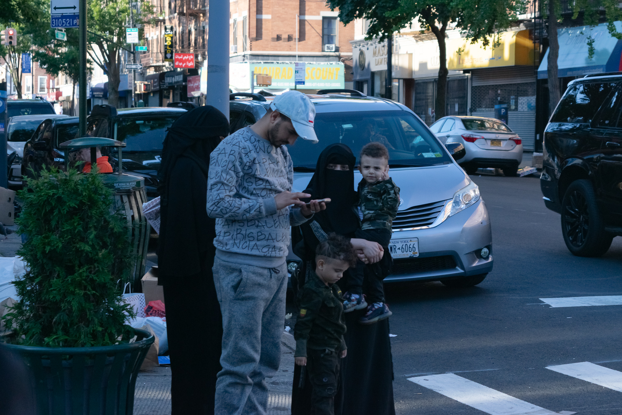 On a Brooklyn sidewalk, a man in a white cap and patterned grey sweatsuit looks down at his phone while a woman in a black niqab holds a toddler in camouflage on her hip; a second small child in matching camo stands beside them, and a silver minivan waits in the street behind.