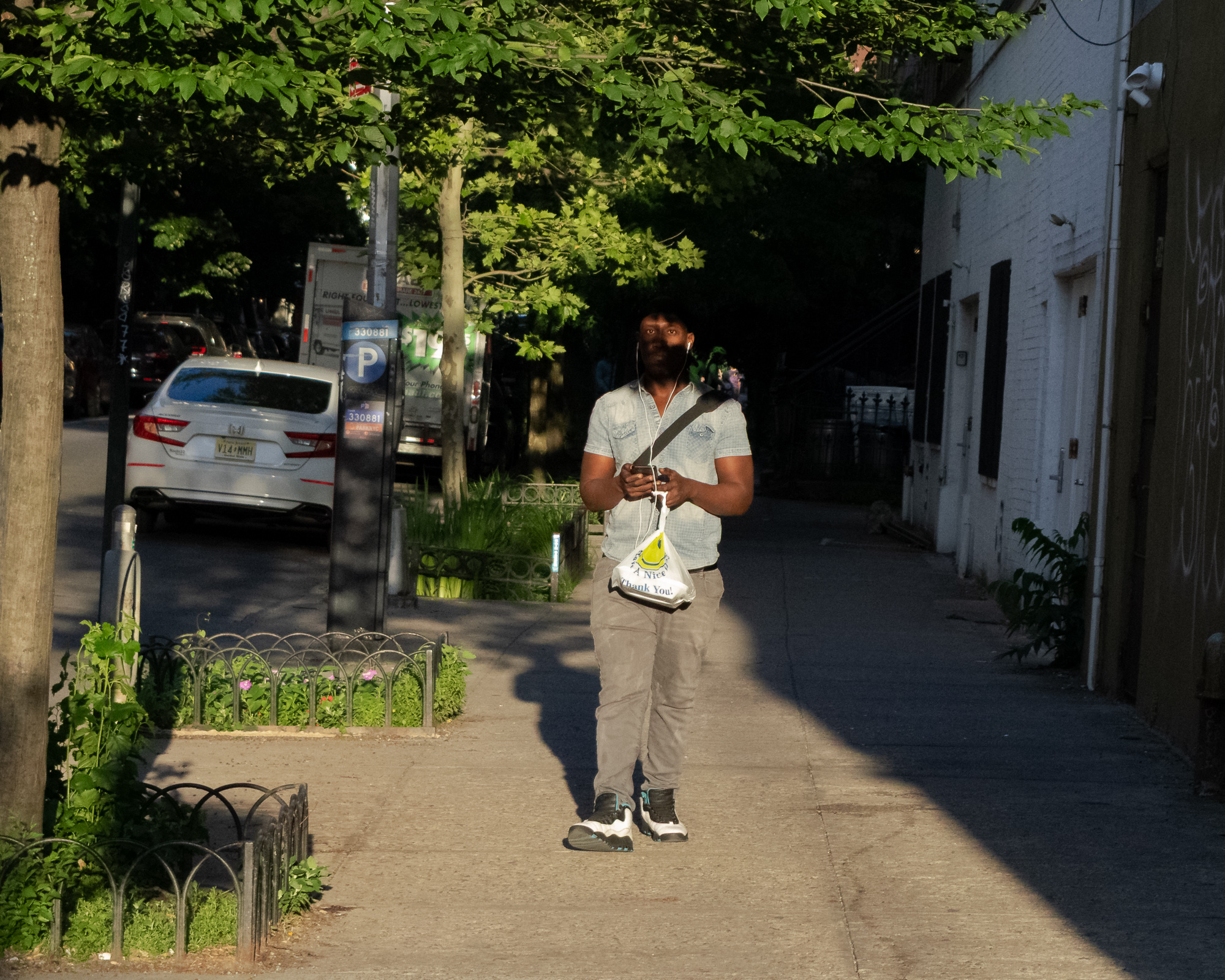A man in a light blue short-sleeve shirt and grey trousers walks toward the camera on a shaded sidewalk, white earbud cords running from his phone and a white plastic bag reading 'Have A Nice Day, Thank You' hanging from his wrist. Behind him a white sedan is parked at the curb beside a Muni-Meter, with sunlit tree canopy overhead and a white brick building to the right.