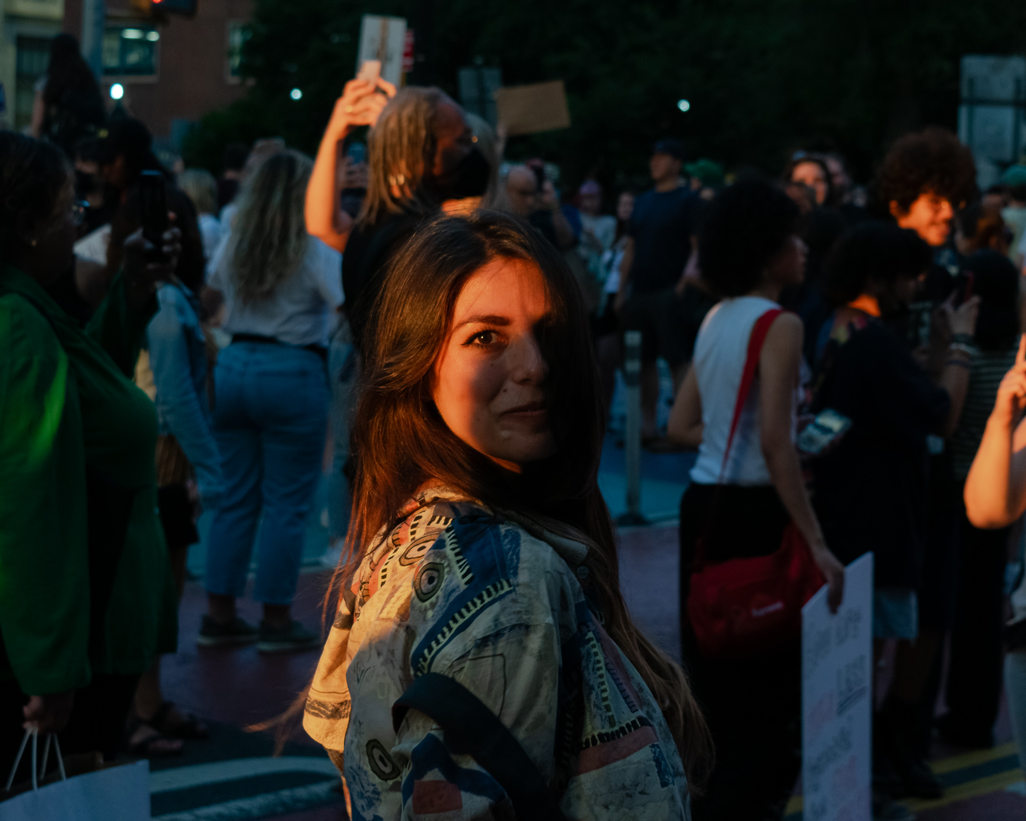 At twilight in a New York City street crowd, a young woman with long brown hair looks back over her shoulder at the camera, her face lit warm orange while the crowd behind her falls into blue shadow. She wears a loose patterned jacket in cream, blue and rust; protesters around her hold cardboard signs and raise phones overhead.