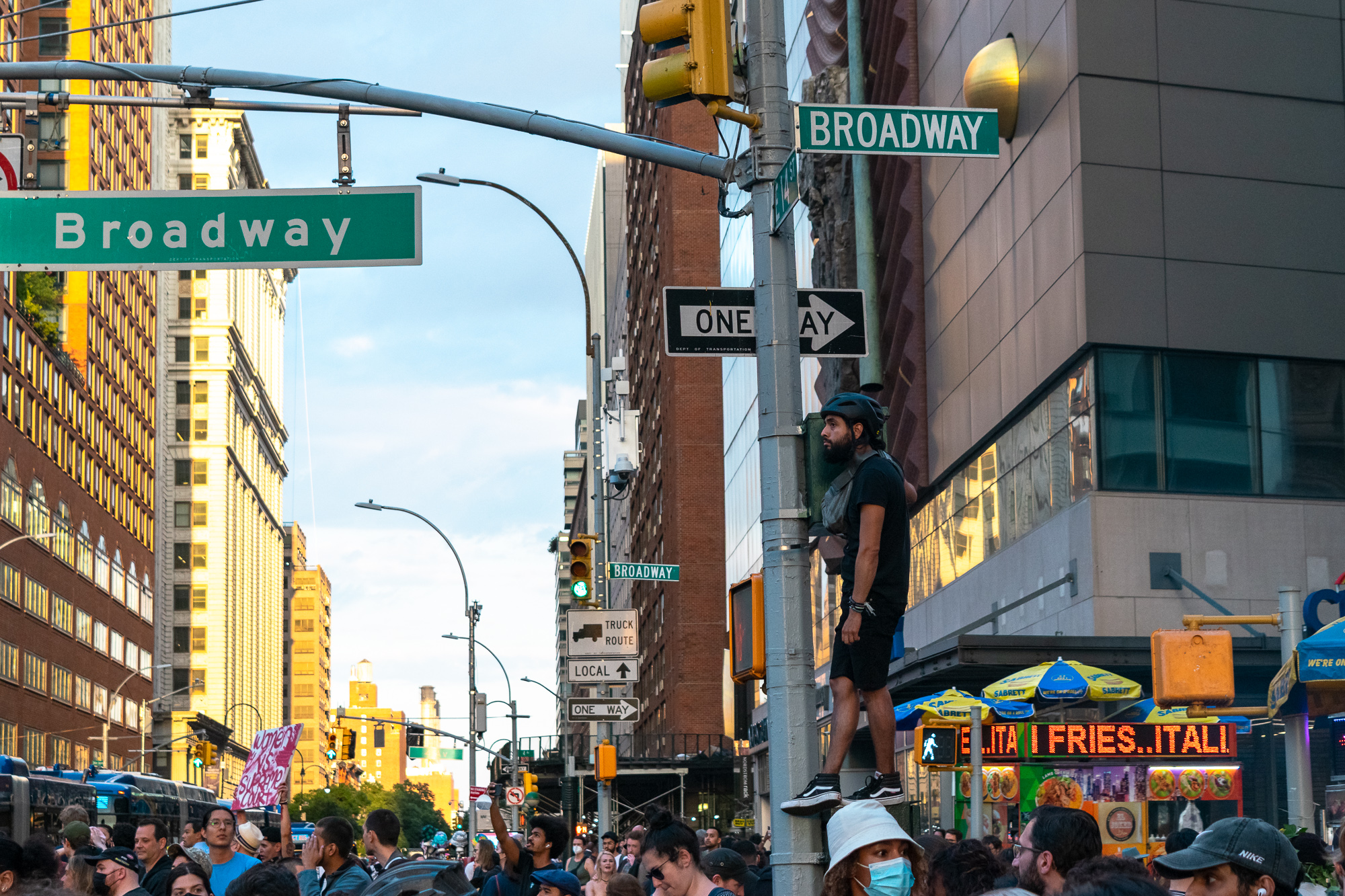 A man in a black t-shirt, shorts and a bicycle helmet stands on a ledge partway up a traffic signal pole at a Broadway intersection, looking off to the side above a dense crowd. Green Broadway street signs, a ONE WAY arrow and a Sabrett hot dog cart with yellow and blue umbrellas fill the frame around him.