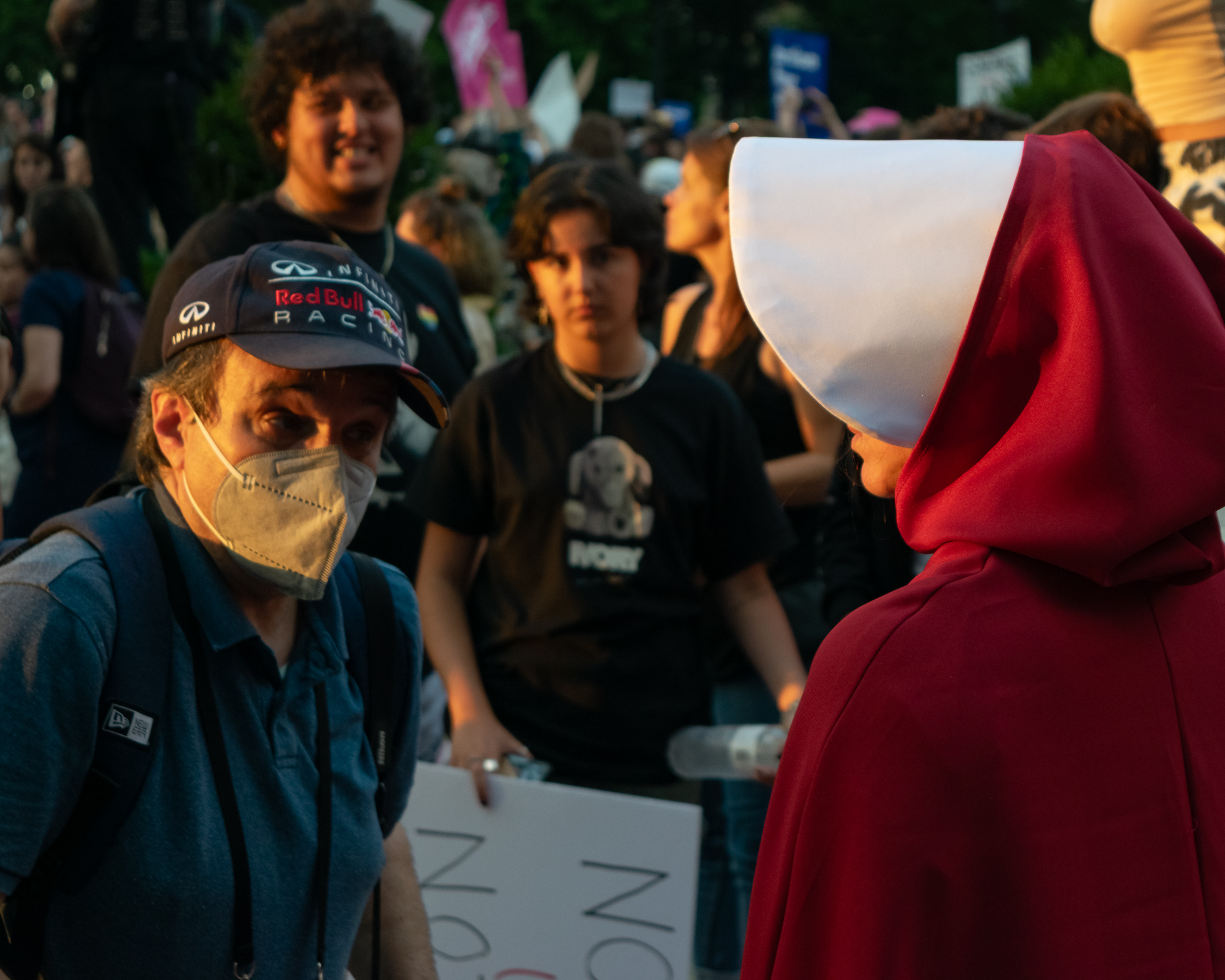 At a crowded evening protest, a man in a navy Infiniti Red Bull Racing cap and a beige KN95 mask turns to look toward a person in a red hooded cloak and white winged bonnet at the right edge of the frame. Behind them, blurred demonstrators in dark t-shirts hold signs in warm low-angle sunlight.