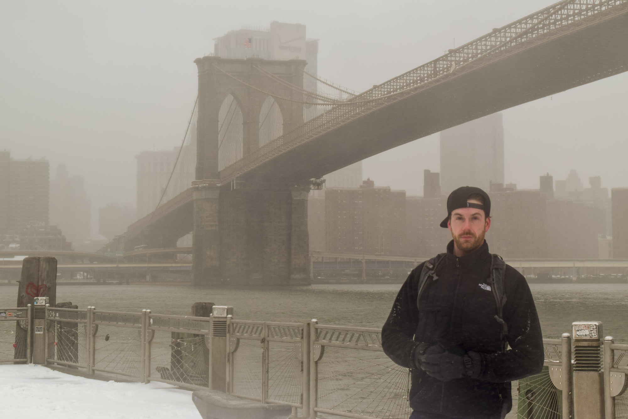 A bearded man in a black backpack, dark jacket and backwards cap stands on a snowy riverside promenade with the Brooklyn Bridge and a fog-obscured Manhattan skyline behind him. Snow clings to his shoulders and gloves, and a metal railing runs along the edge of the East River.