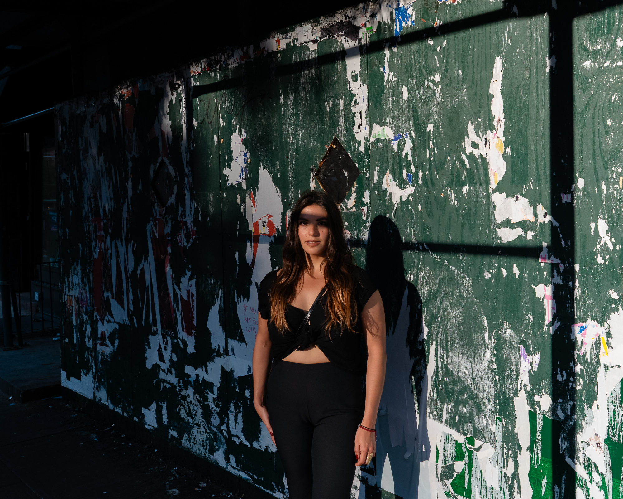 A young woman with long brown hair stands in late golden sunlight against a green plywood construction wall covered in shredded poster remnants, wearing a black knotted top and black leggings with a thin crossbody strap. A hard-edged shadow cuts diagonally across her eyes and throws her silhouette onto the wall beside her.