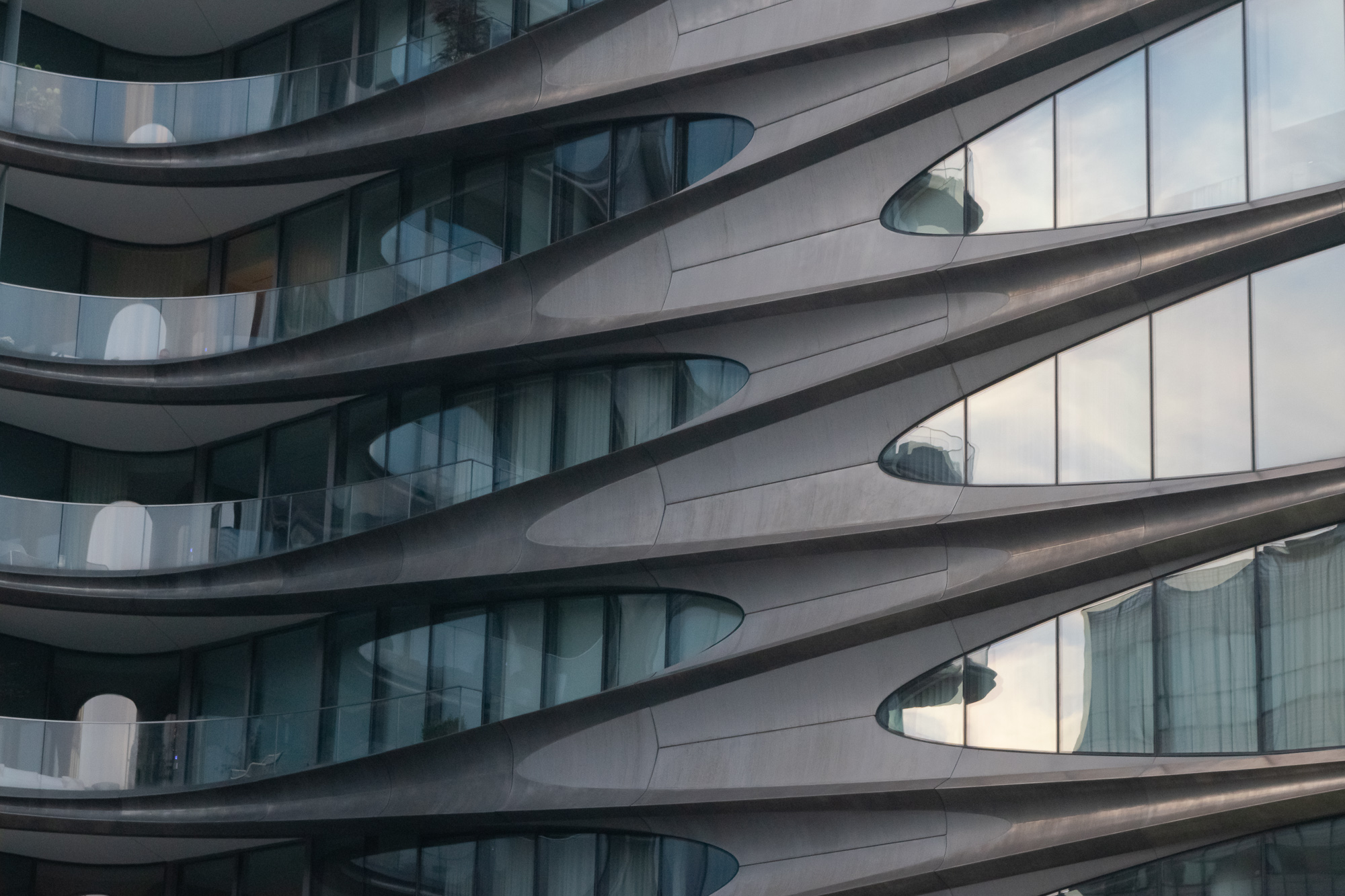 Close-up of a modern apartment building facade made of interlocking curved metal bands that form pointed chevron shapes, with recessed glass balconies and windows reflecting pale evening sky between them.