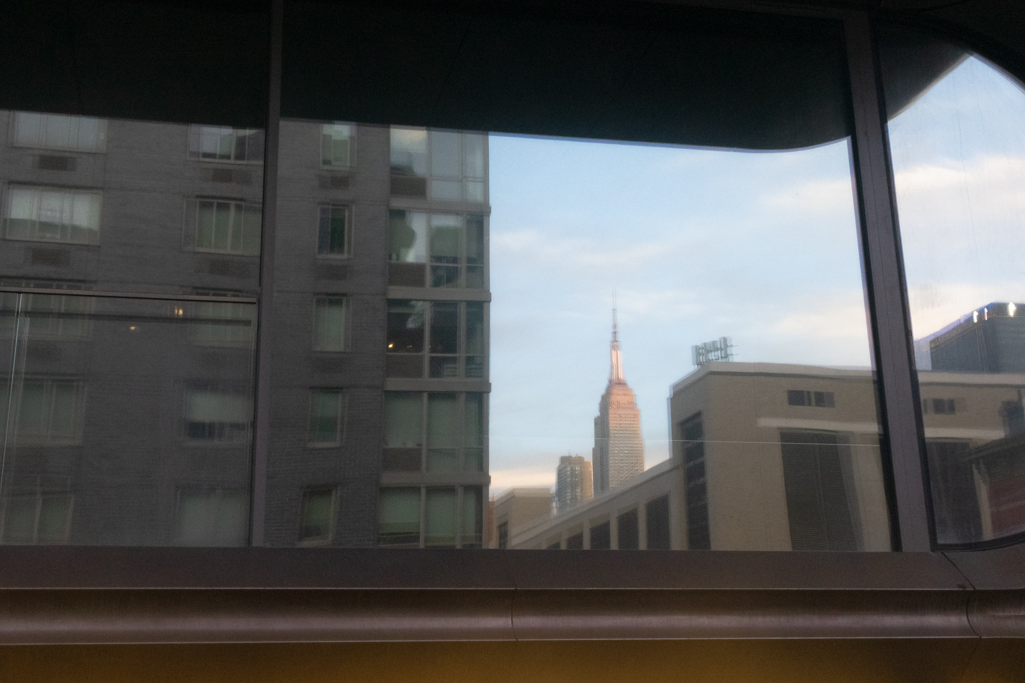 View through a large tinted window with a dark upper frame: at left a gray brick and glass apartment tower fills the frame, and in a gap of pale evening sky at center the upper floors and spire of the Empire State Building catch warm light, with low rooftops and a rooftop antenna array to the right.