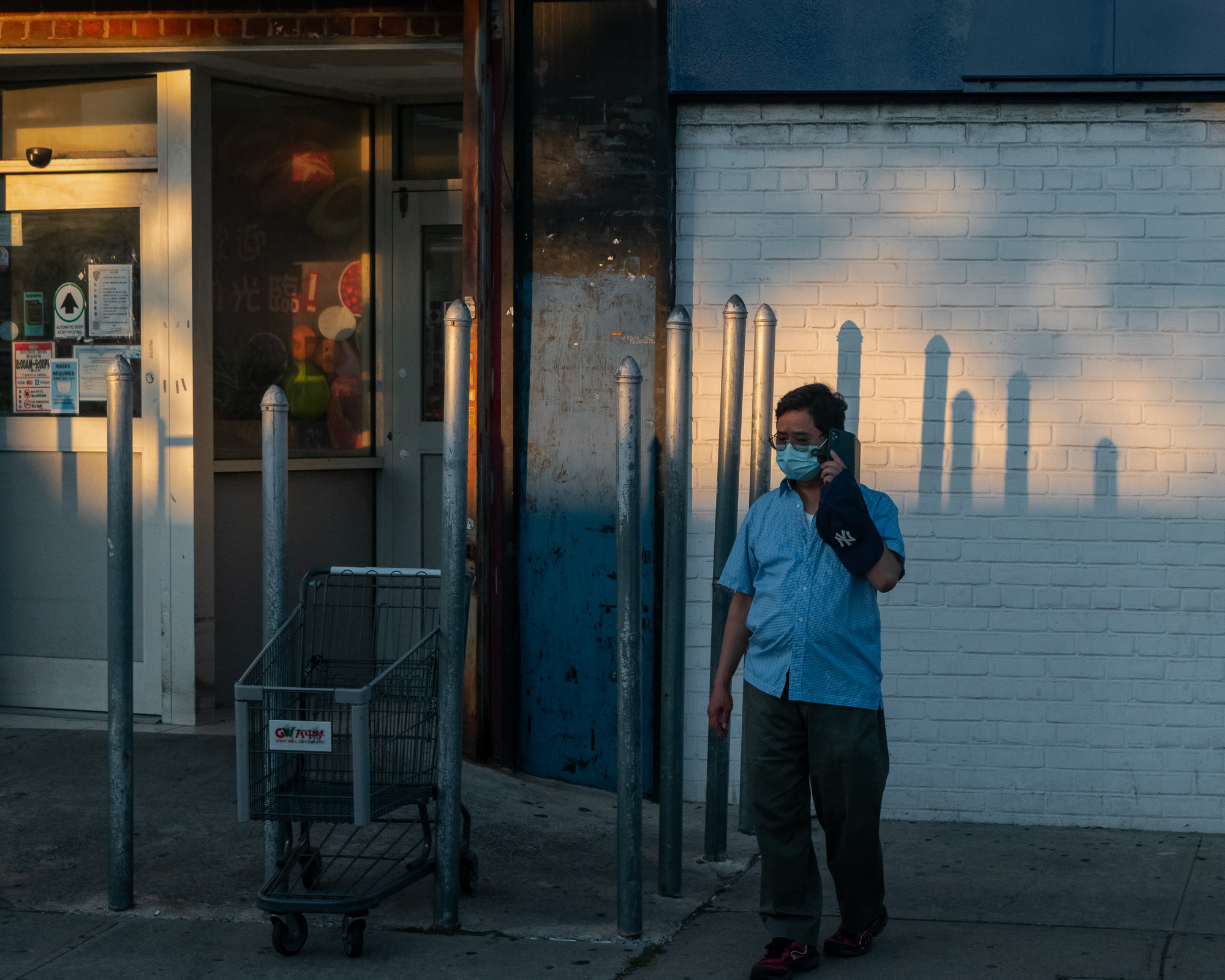 A man in a light blue checked shirt and surgical mask holds a phone to his ear with a navy Yankees cap in his hand, standing on a sidewalk in front of a white painted brick wall where low sun casts long shadows of steel bollards. To his left, an empty Great Wall Supermarket shopping cart sits outside the store's glass entrance.