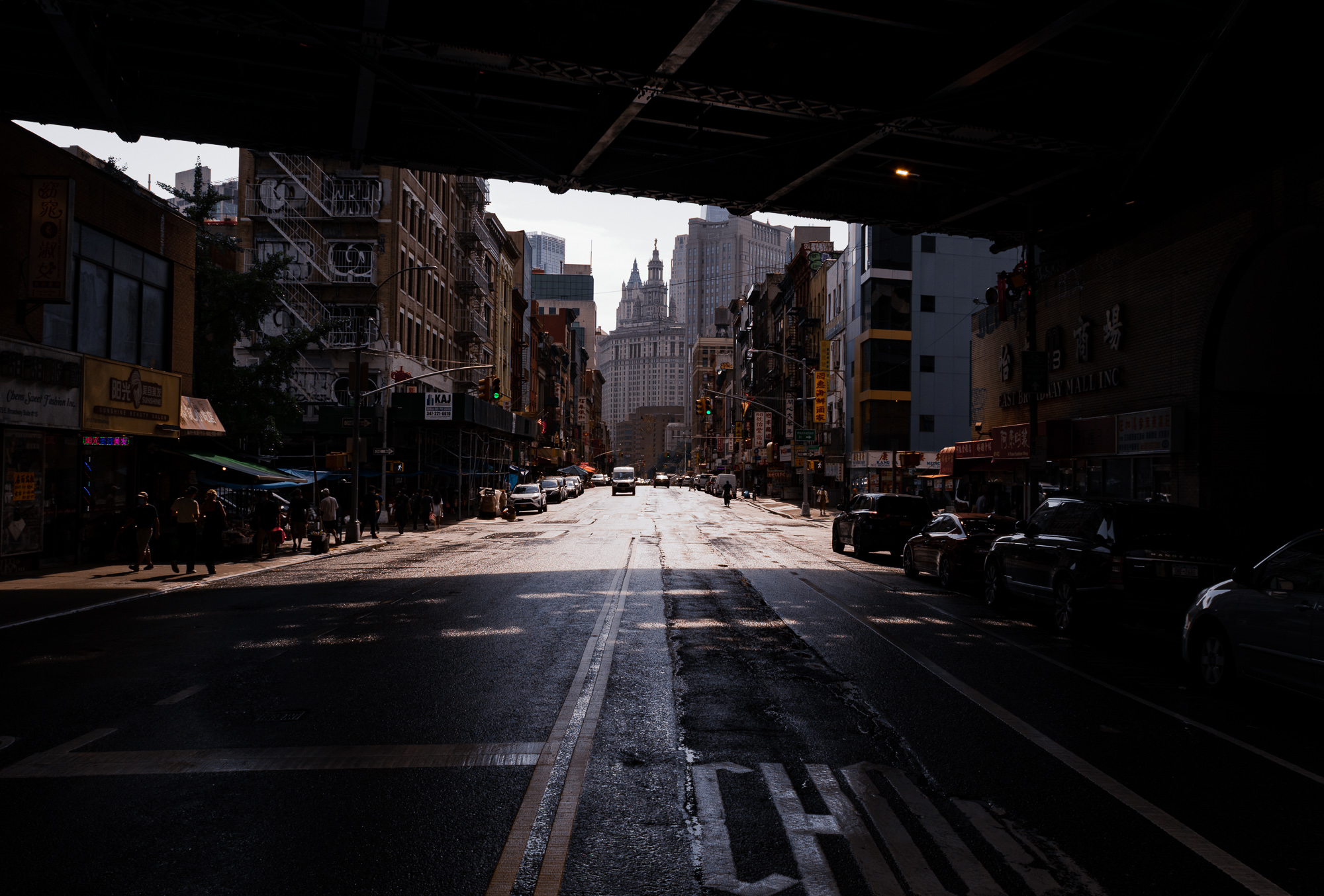 View from beneath a dark steel bridge overpass looking up a wide Chinatown street in Manhattan, with Chinese storefront signs, parked cars and pedestrians on the left sidewalk, and the twin-towered Municipal Building rising in hazy light at the end of the avenue.