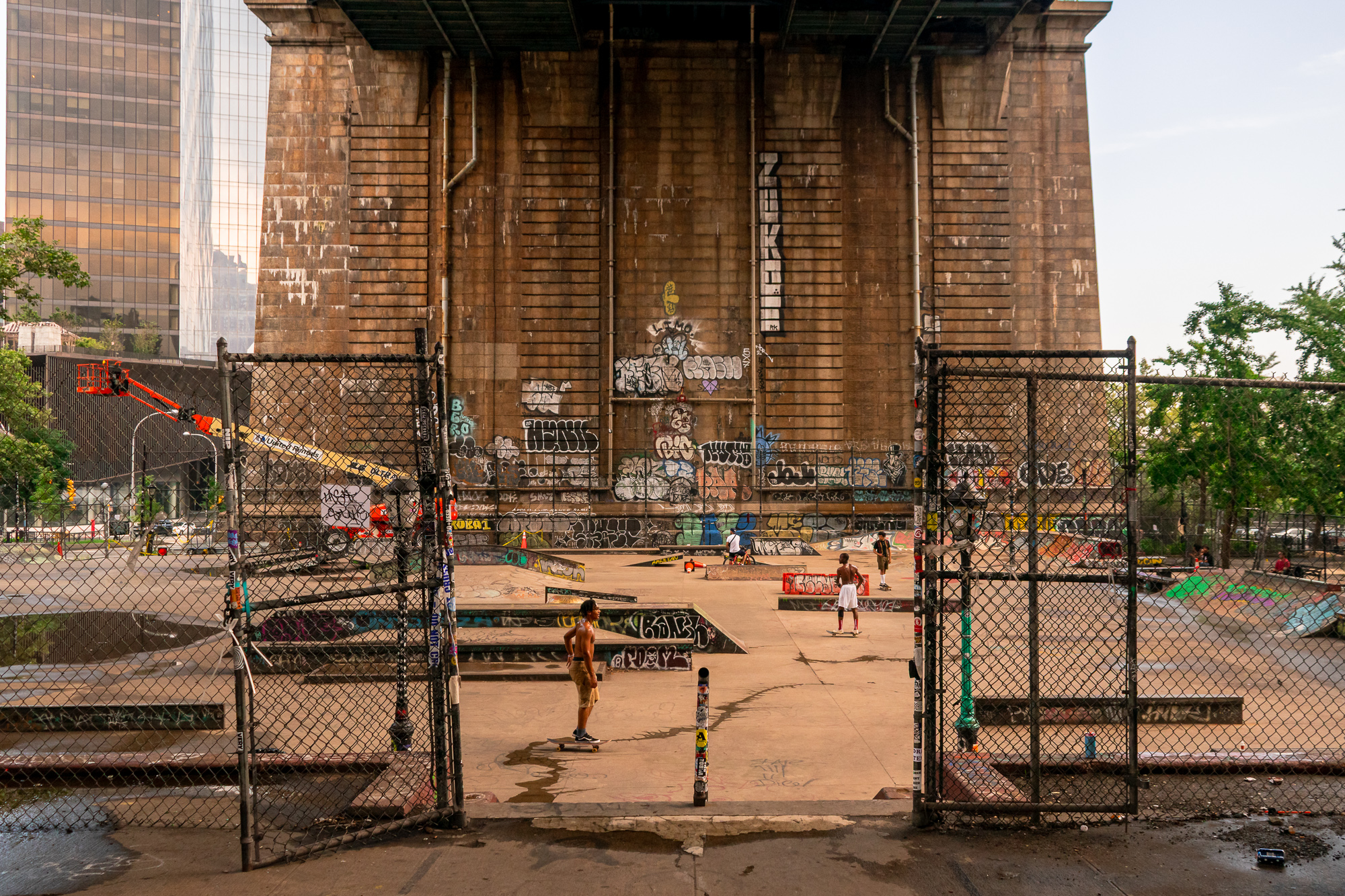 View through an open chain-link gate into a concrete skatepark beneath the massive graffiti-covered stone anchorage of the Brooklyn Bridge, where a shirtless skater rolls toward the camera and others ride ramps in the background. An orange boom lift and glass office towers rise behind the park at left.