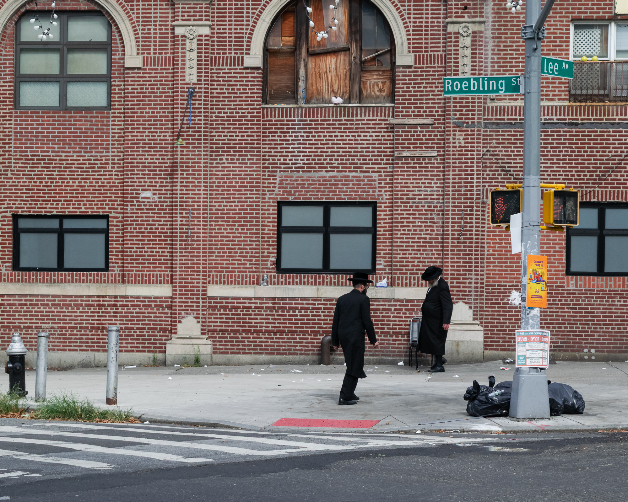 On a Brooklyn corner, one man in a black coat and wide-brimmed hat walks across the sidewalk while an older bearded man in similar dress stands still beside a folding chair. Behind them rises a red brick synagogue wall with arched windows and boarded wooden panels; green Roebling St and Lee Av signs and a crosswalk signal hang from a pole at right.