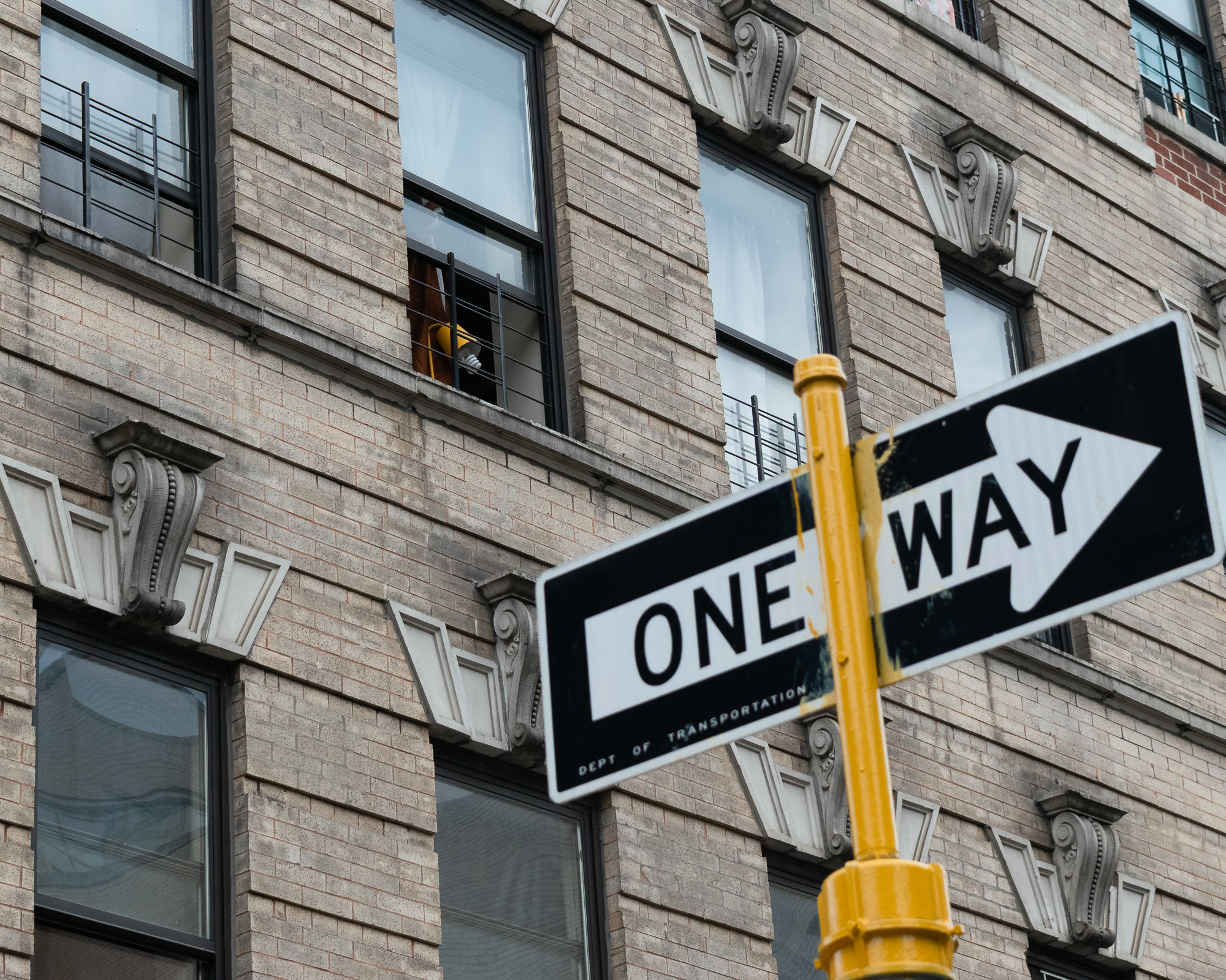 A black and white ONE WAY street sign mounted on a yellow pole, tilted diagonally in front of a tan brick apartment building with carved stone brackets under the windows. Yellow paint has dripped down the sign where it meets the pole, and a yellow lamp is visible through an open second-floor window.