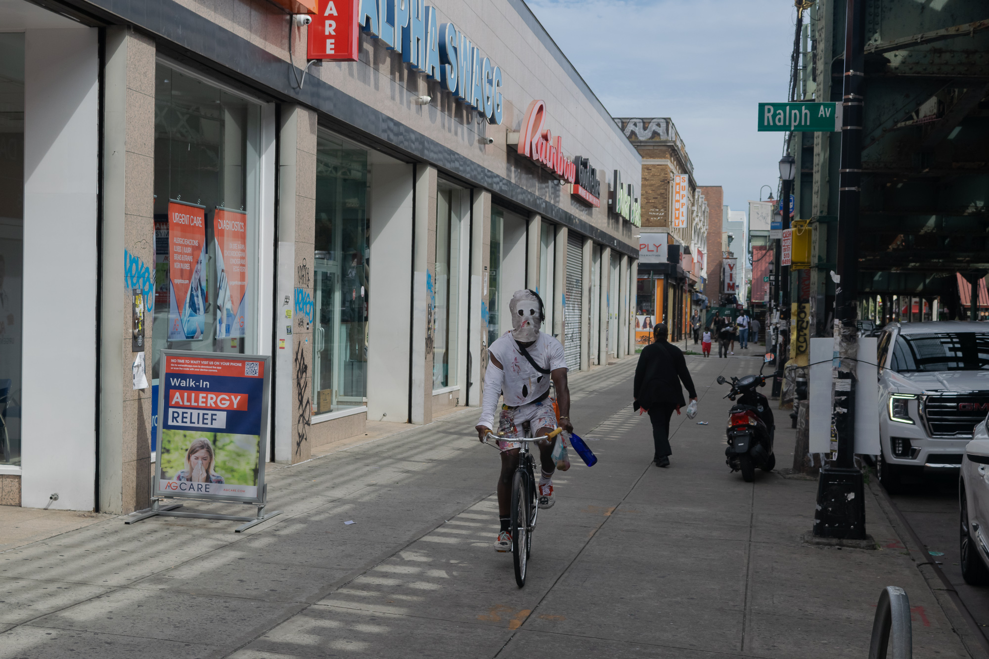 A cyclist wearing a paint-spattered white hood over the entire head and a matching torn shirt rides along a Brooklyn sidewalk past shuttered storefronts, carrying a blue bottle. A Walk-In Allergy Relief sandwich board stands to the left and the green Ralph Av street sign hangs under the elevated tracks at right.