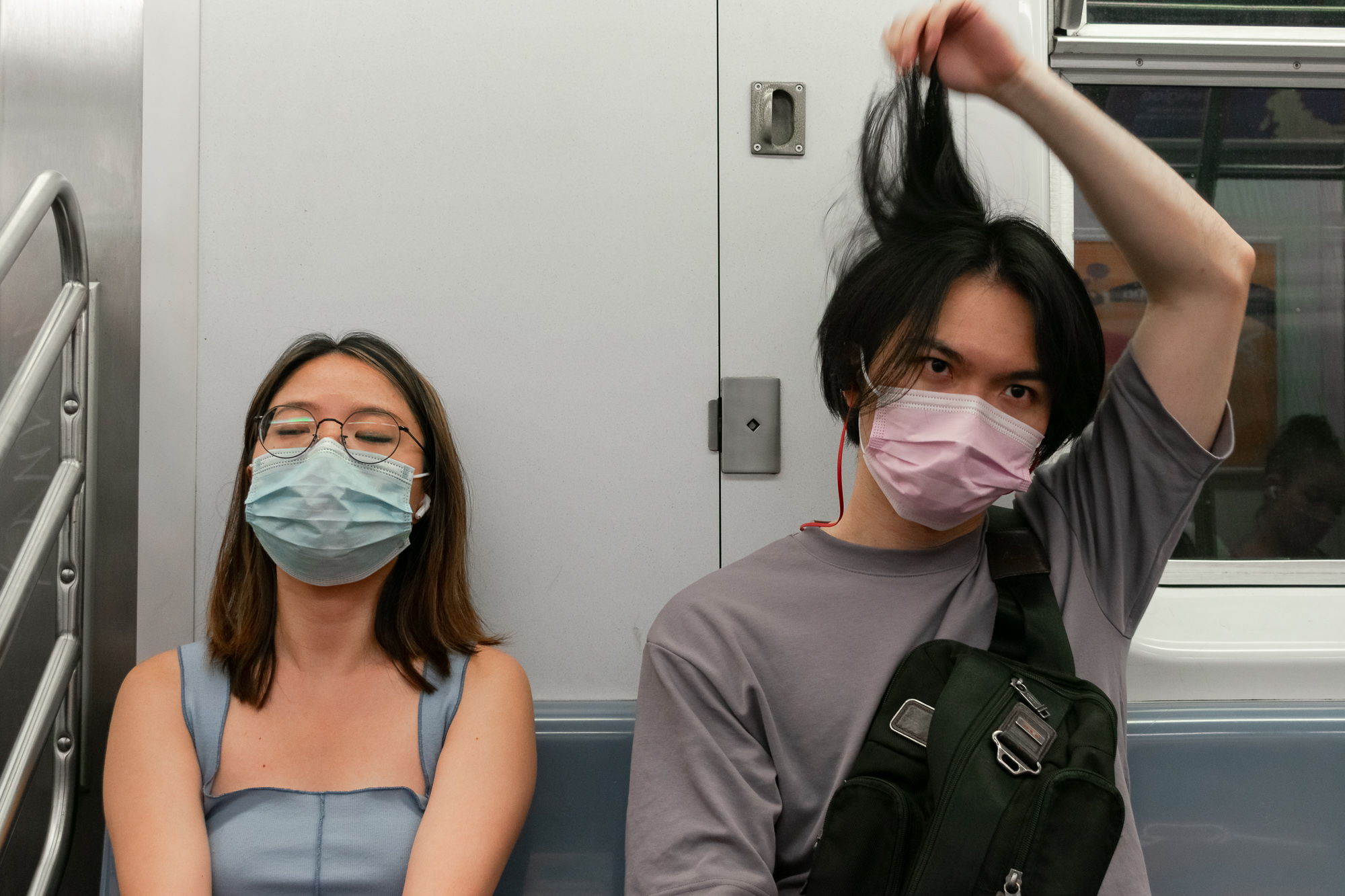 Two masked passengers sit side by side on a New York subway car. On the left, a woman in a light blue tank top and round glasses rests with her eyes closed behind a surgical mask; on the right, a young man in a gray sweatshirt with a black sling bag holds a fistful of his own long dark hair straight up above his head, eyes fixed on the camera.
