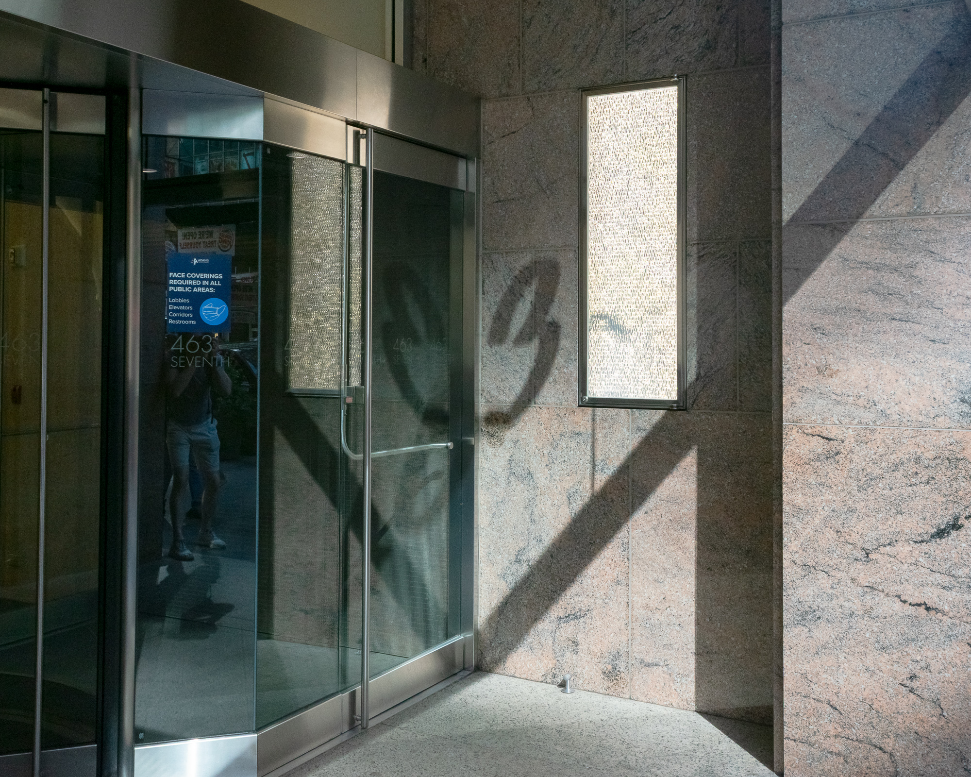 A glass and steel office building entrance beside a pink-and-grey granite wall, where a hard diagonal shadow cuts across the stone and a tall vertical light panel glows in its frame. A blue sign on the glass reads 'Face coverings required in all public areas,' and the photographer's reflection is visible in shorts and sneakers.
