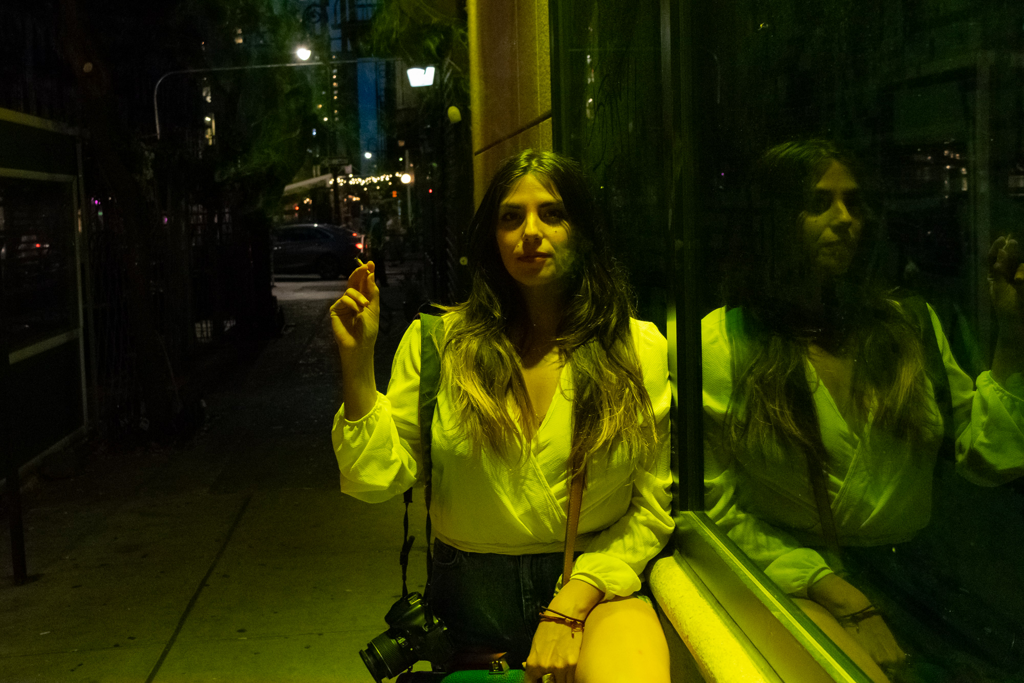 At night on a New York sidewalk, a woman with long dark hair in a white wrap blouse and jeans sits against a plate glass storefront, holding a cigarette up near her shoulder, a DSLR camera hanging at her hip. Green and yellow light from the window washes over her and throws her mirrored reflection across the glass to the right.