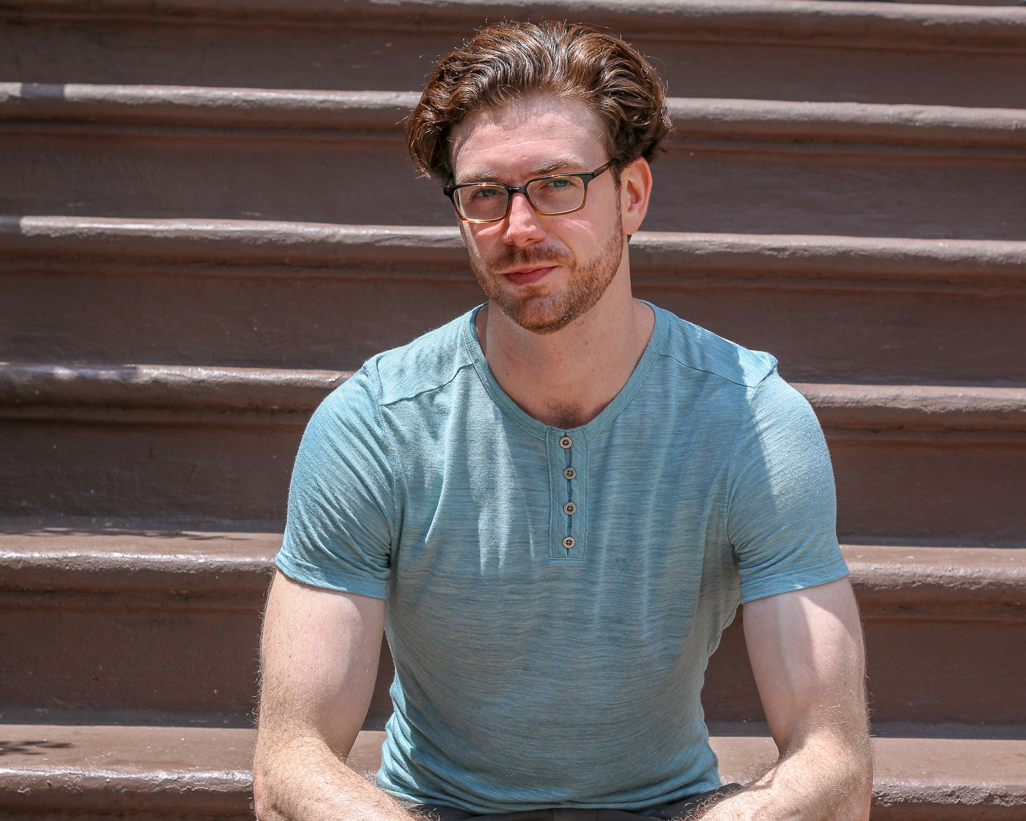 A bearded man with wavy auburn hair and tortoiseshell glasses sits on wide painted brownstone steps in bright midday sun, wearing a teal heathered henley T-shirt with four buttons open at the collar. He faces the camera with a slight smile, arms resting forward, the stoop's brown risers filling the frame behind him.