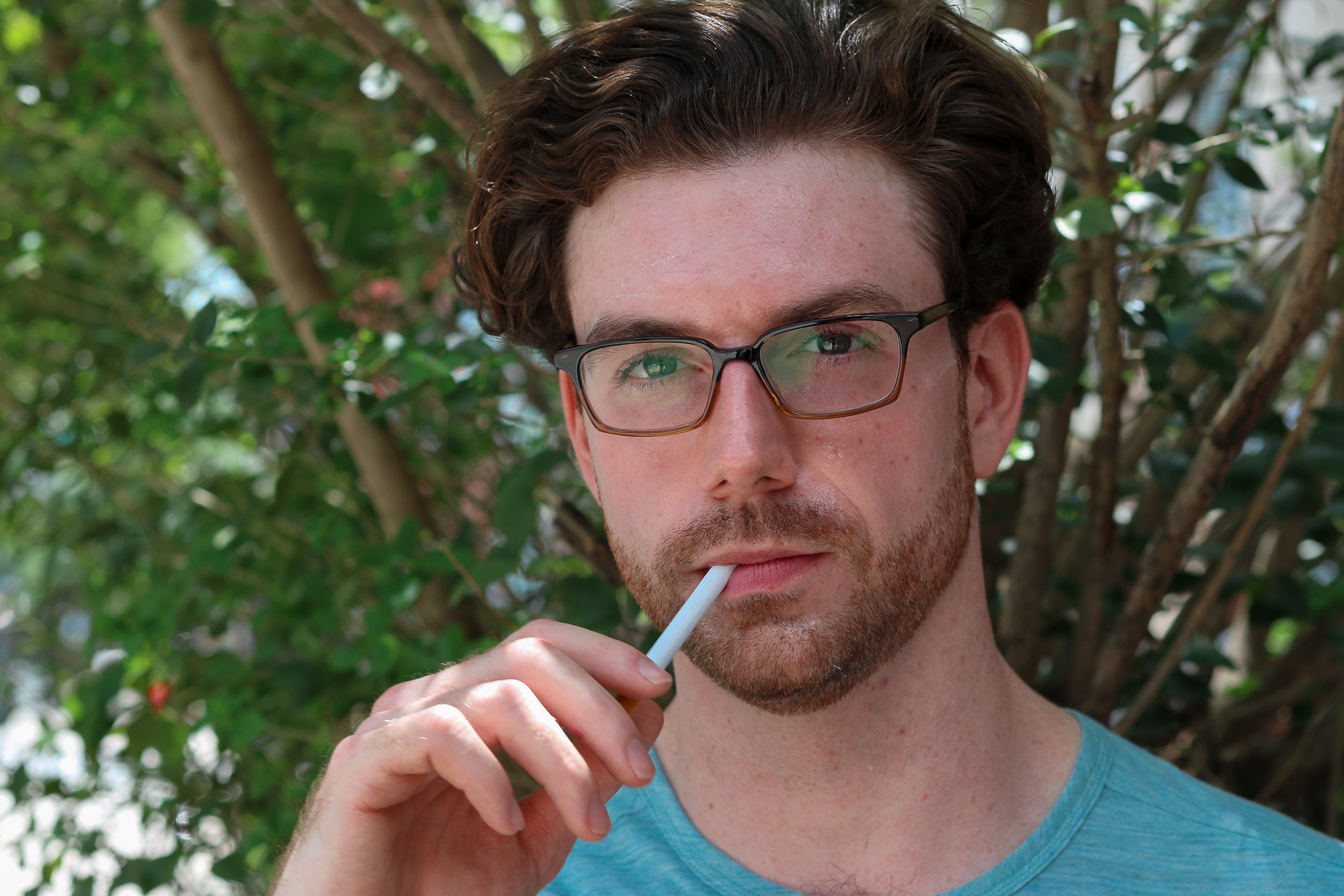 Close-up daylight portrait of a young man with wavy brown hair, a reddish beard and rectangular tortoiseshell glasses, holding a white paper straw between his lips with his right hand. Sunlit green leaves and thin tree trunks fill the background, and he wears a light blue heathered t-shirt.