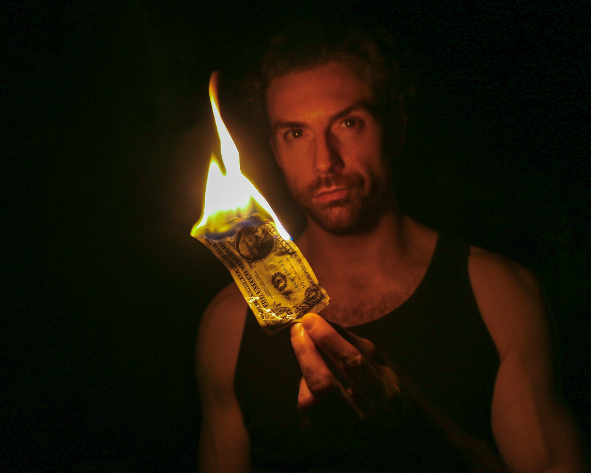 In near-total darkness, a bearded man in a black tank top holds a one dollar bill between his fingers as it burns, the flame rising in a tall yellow and blue spike. The firelight is the only illumination, catching his face, bare shoulder and the serial number still legible on the scorched note.