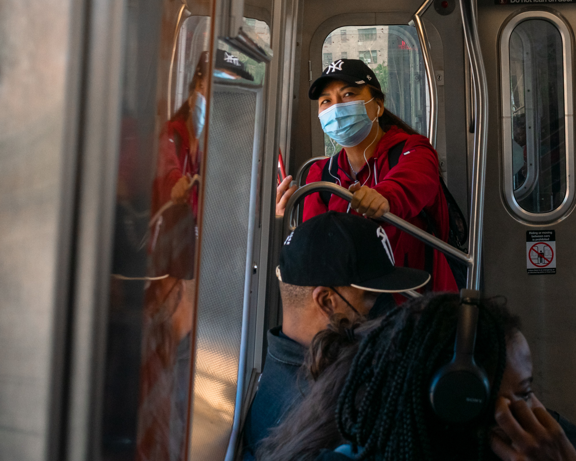 A woman in a red hooded sweatshirt and a black New York Yankees cap wears a blue surgical mask and white wired earphones, holding a steel grab rail near the end-door of a subway car. In the foreground, out of focus, a man in a black cap and a rider in braids with over-ear headphones sit below her, and a reflection of the red jacket repeats in the door glass at left.