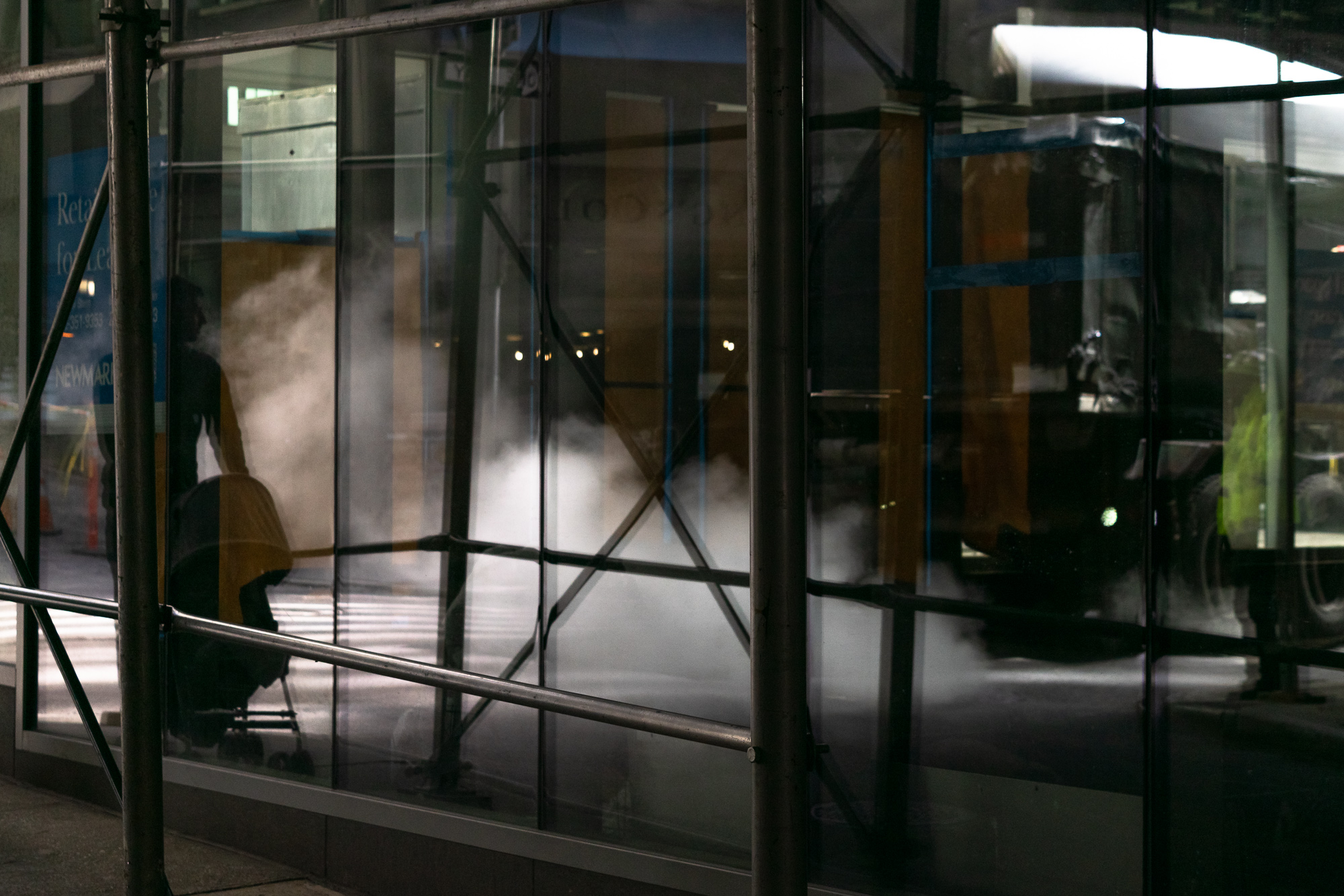 Night view through a scaffolded glass storefront where a seated figure in silhouette sits beside a stroller, with a plume of white steam rising in the street and layered reflections of traffic, traffic cones, and a leasing sign doubling across the panes.