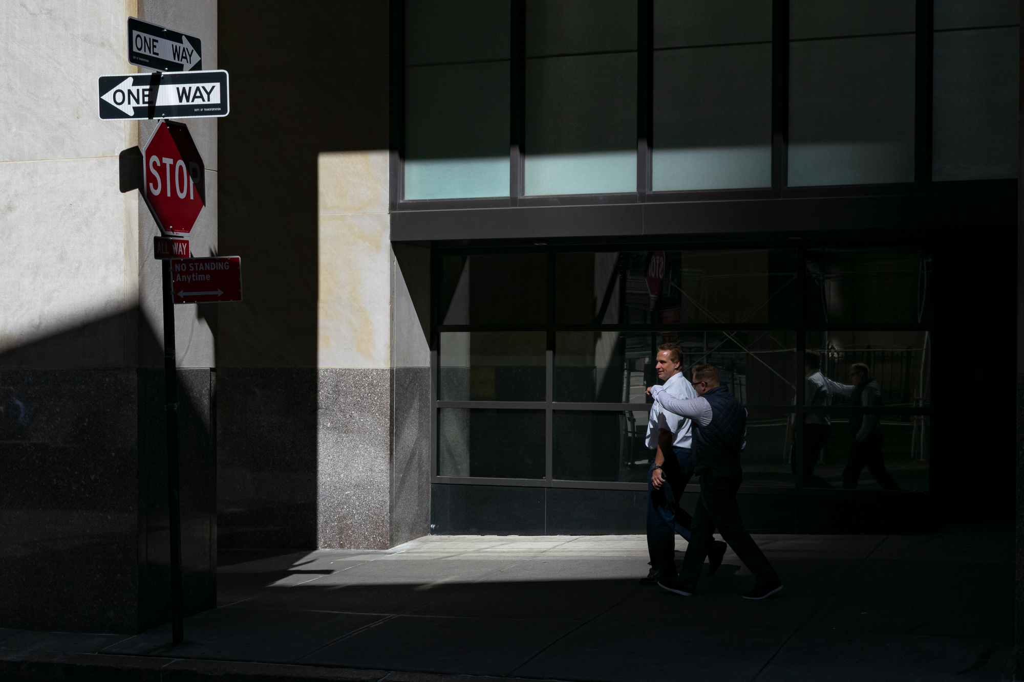 Two men in business casual clothes walk along a sunlit sidewalk past a dark glass office facade, their bodies half-swallowed by deep shadow. At left, a pole carries two One Way arrows, an all-way Stop sign, and a No Standing Anytime plate, and the Stop sign appears again as a reflection in the building's windows.