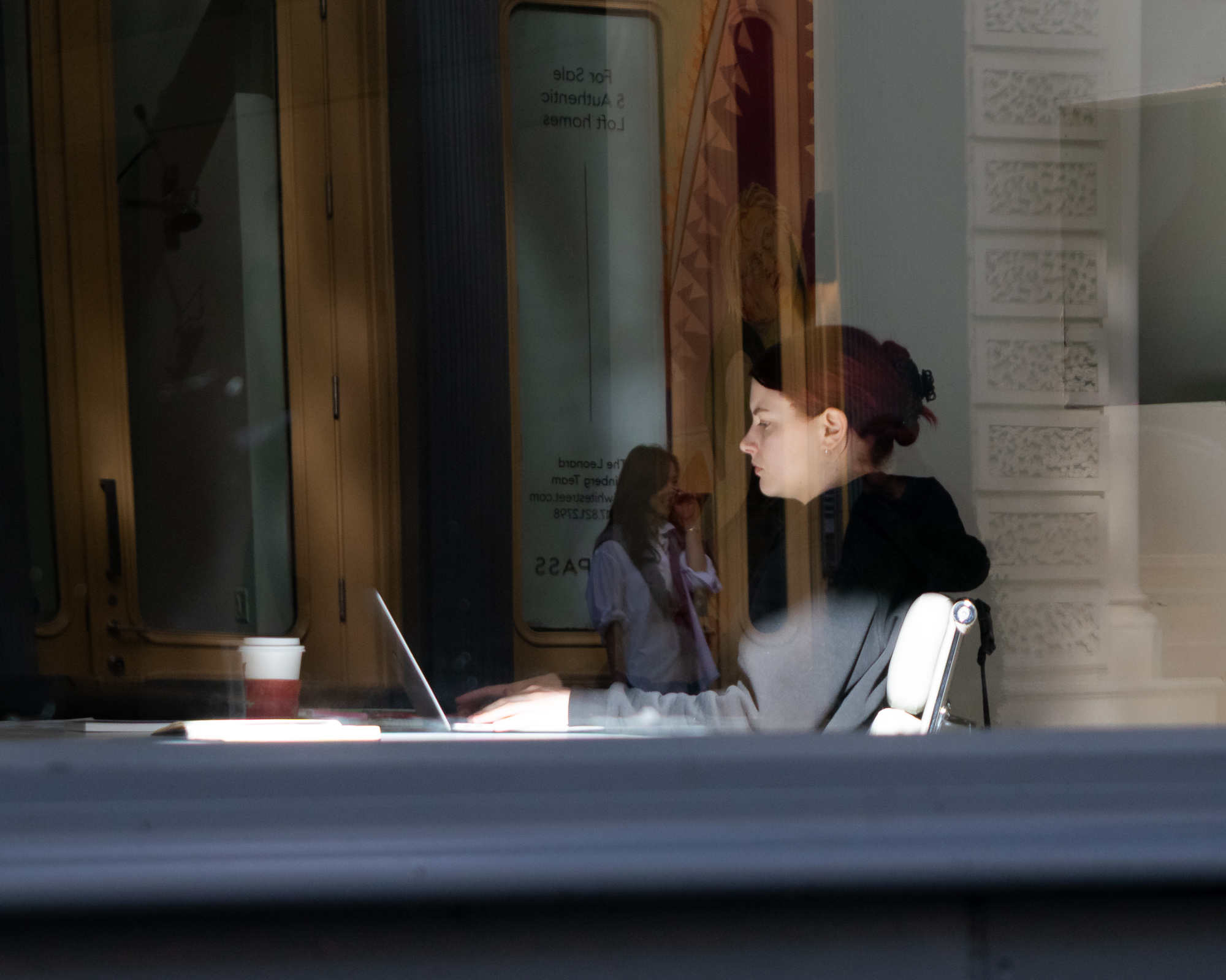 Seen through a large storefront window, a woman with red hair pinned up sits in profile at a white chair, working on a laptop beside a takeout coffee cup, while the glass reflects a second woman in a pale blouse standing outside and the carved white facade of a building across the street.