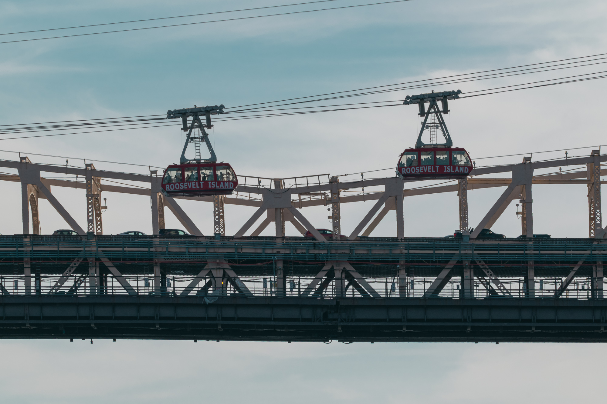 Two red aerial tram cabins lettered ROOSEVELT ISLAND hang from cables as they pass each other above the steel truss and traffic deck of the Queensboro Bridge, with overhead wires crossing a pale overcast sky.
