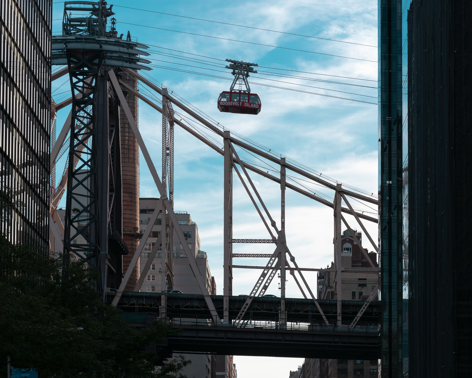 A red Roosevelt Island tram cabin hangs from its cables in a blue sky above the steel trusses and stone tower of the Queensboro Bridge, framed on both sides by dark glass office towers on East 59th Street.