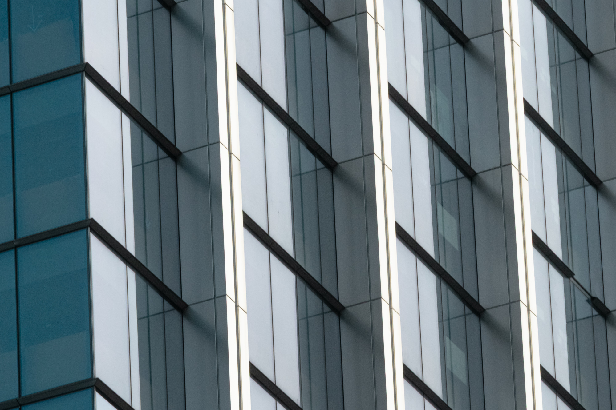 Tight telephoto view of a glass office tower facade, where vertical white mullions and dark spandrel bands cut diagonally across gray-blue reflective panels. At the left edge, a section of deeper teal glass meets the corner of the building.