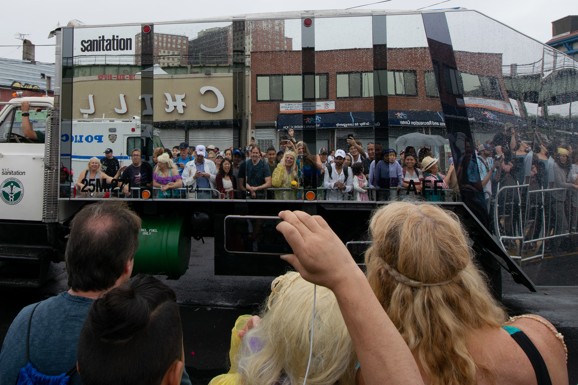 A New York City sanitation truck clad in mirrored panels reflects a rain-soaked crowd pressed against metal barricades along a Coney Island street, with reversed storefront signs and a police van visible in the reflection. In the foreground, spectators face away from the camera, one raising a phone to photograph the passing truck.