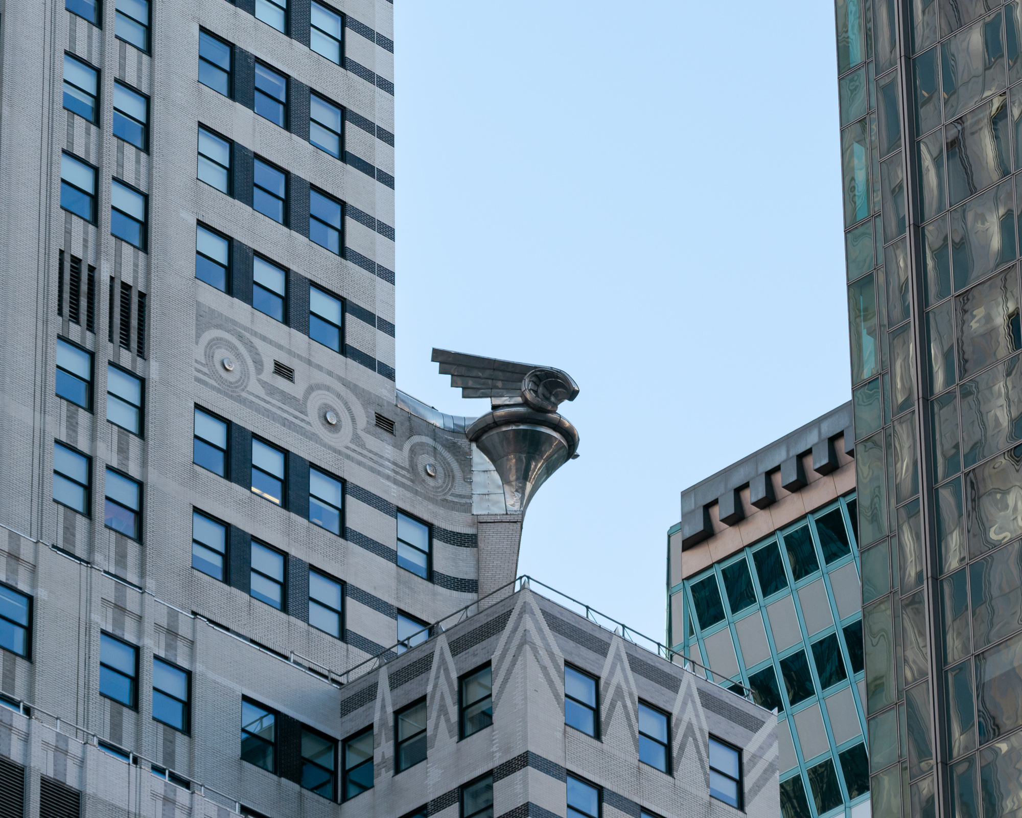 A steel eagle gargoyle projects from the corner of a pale brick Art Deco tower, set against clear blue sky, with a band of hubcap-motif brickwork and chevron patterns below and a green-tinted glass building to the right.