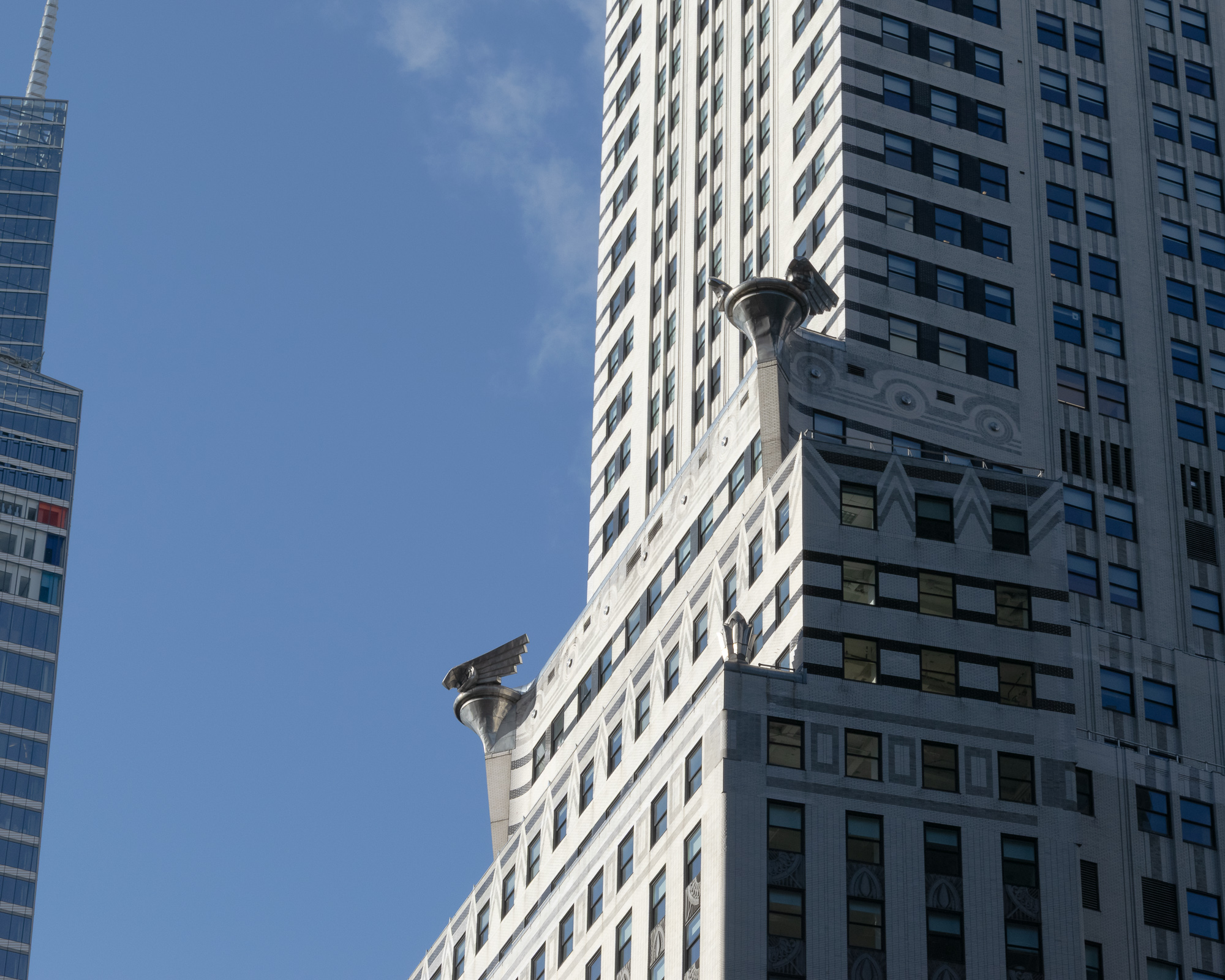 Detail of the Chrysler Building's upper setbacks in Manhattan, with two stainless steel eagle gargoyles projecting from the white brick corners against a clear blue sky. A glass tower with a thin spire rises at the far left edge of the frame.