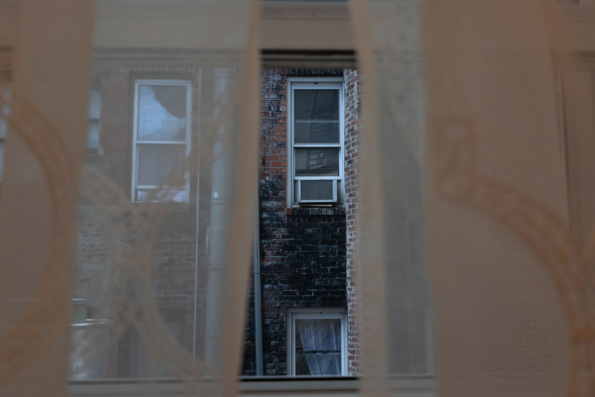 A narrow vertical gap between blurred plywood construction panels reveals a soot-darkened brick facade across an airshaft, with a white-framed sash window holding a window air conditioner above a lower window screened by a pale curtain.