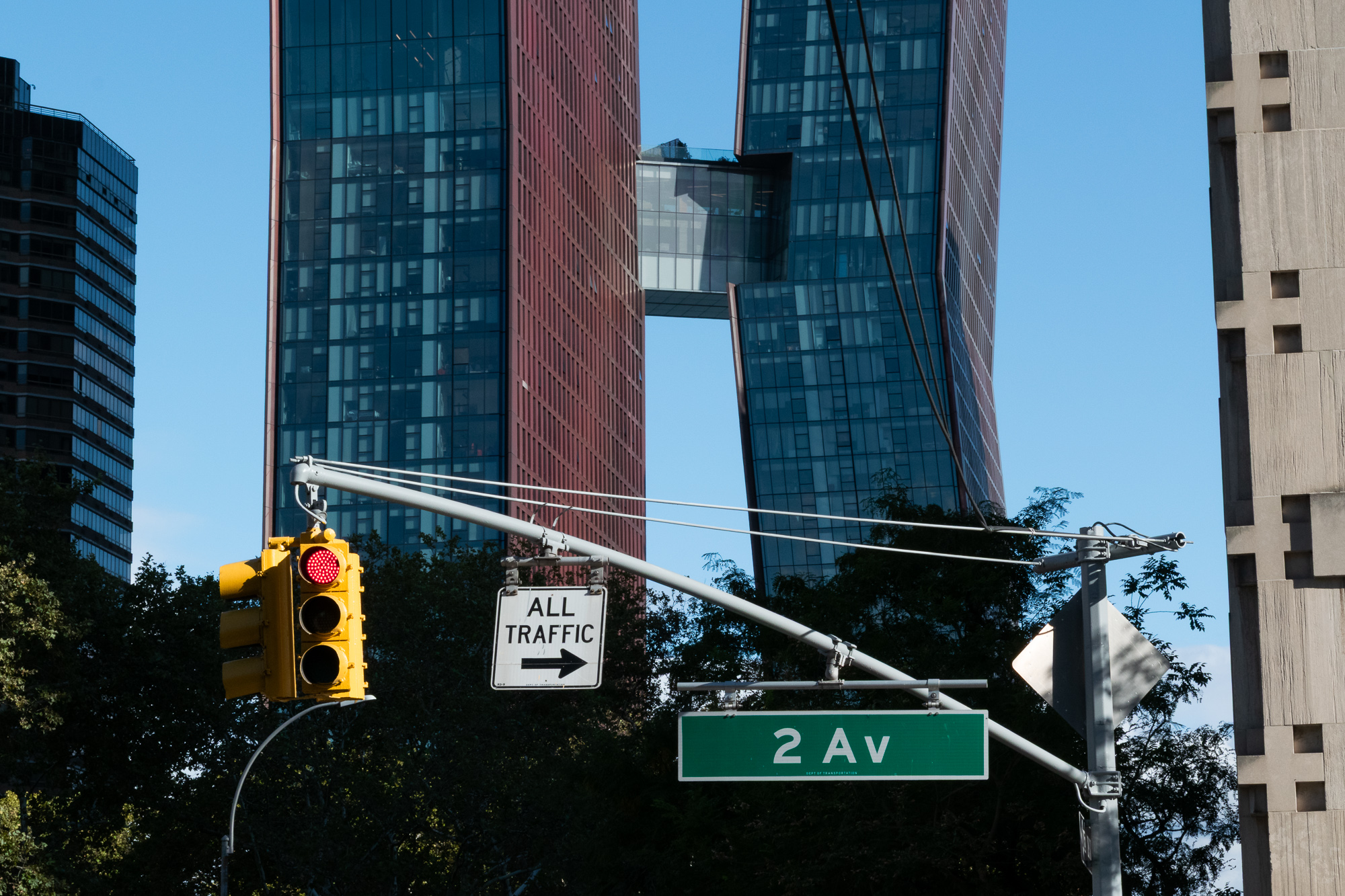 A yellow New York traffic signal glowing red hangs from a gray mast arm alongside a white 'ALL TRAFFIC' sign with a right arrow and a green '2 Av' street sign. Behind them, two leaning glass-and-copper towers joined by a high skybridge rise above a line of trees into a clear blue sky.
