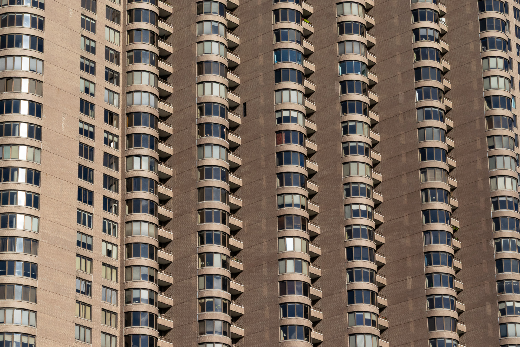 Tight telephoto view of a tan and brown brick apartment tower filling the frame, with vertical columns of rounded balcony bays and dark tinted windows repeating floor after floor. A small potted plant sits on one upper balcony and blinds are drawn at different heights across the glass.