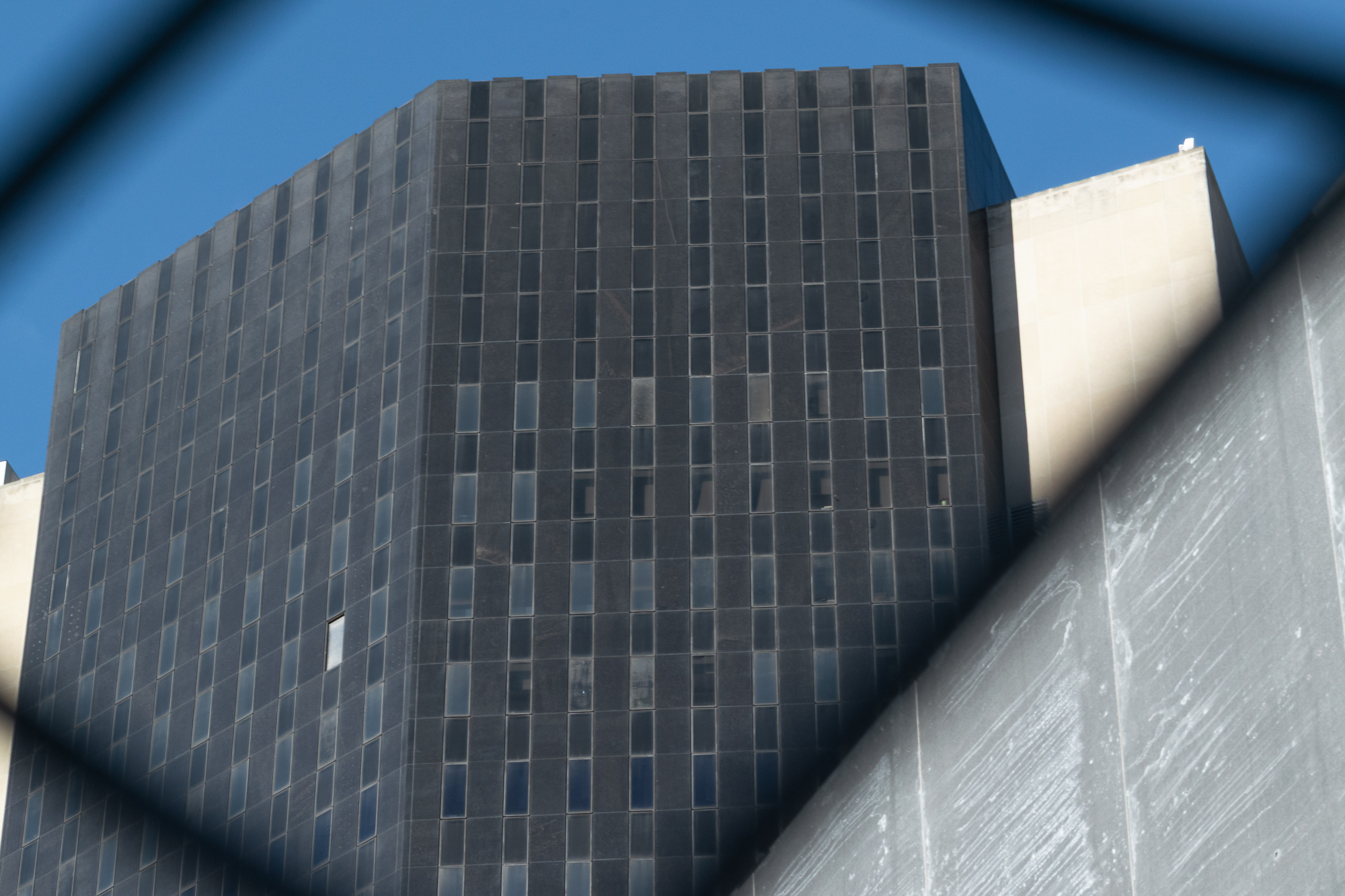 Looking up through the diamond gap of a chain-link fence at the corner of a dark granite-clad office tower with narrow vertical windows set against a deep blue sky. A pale limestone building rises behind it on the right, and a stained concrete wall cuts diagonally across the lower right of the frame.