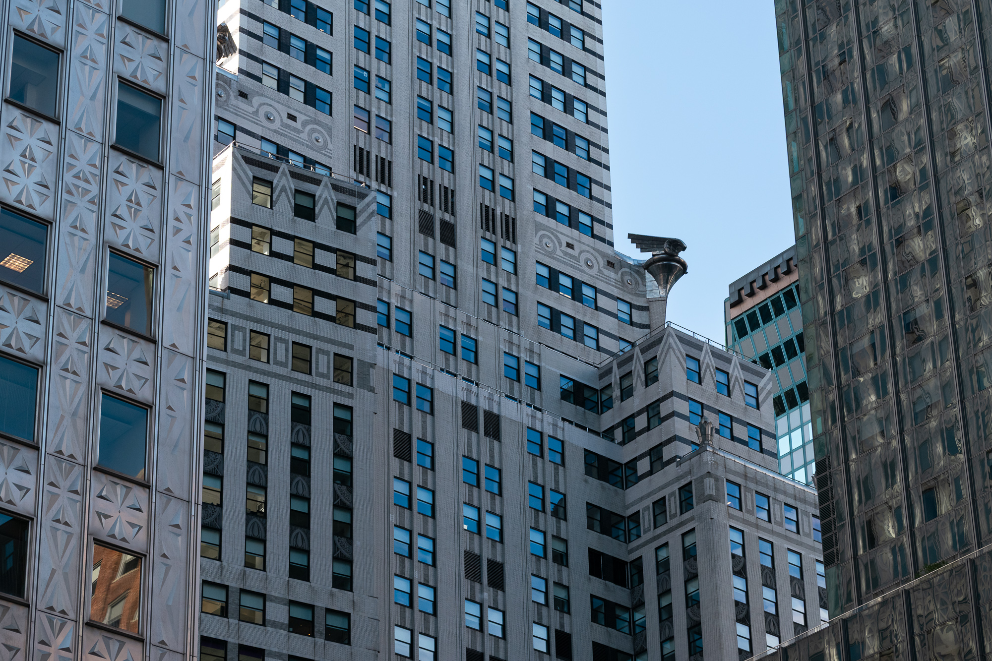 A tight telephoto view of the Chrysler Building's brick and steel setbacks, with a chrome eagle gargoyle projecting from a corner near the center, framed on the left by a patterned stone facade and on the right by a mirrored glass tower reflecting rippled shapes.