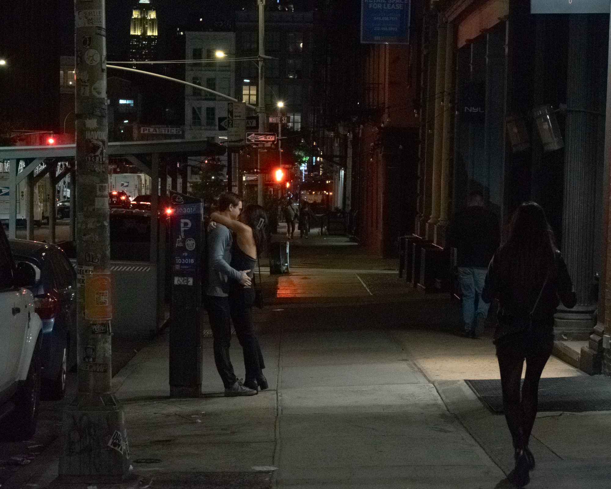 At night on a Tribeca sidewalk, a man in a gray long-sleeve shirt and a woman in a dark off-shoulder top stand pressed together in an embrace beside a blue parking payment kiosk, lit by a warm pool of streetlight. A woman in a dark coat walks past in the foreground at right, and a green WHITE ST sign and a red traffic signal glow further down the block.