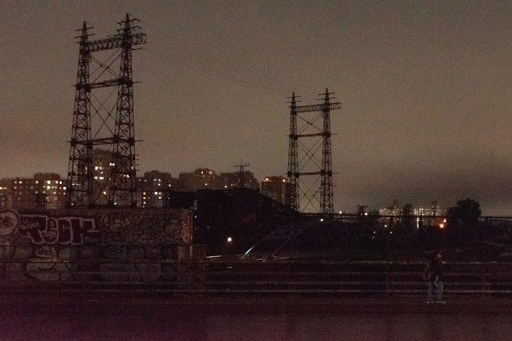 Two tall lattice electrical transmission towers stand in silhouette against a brown-orange night sky, with lit apartment blocks glowing behind them. Below runs a long graffiti-covered concrete wall, a dark embankment, and a railing where a single figure stands at the lower right.
