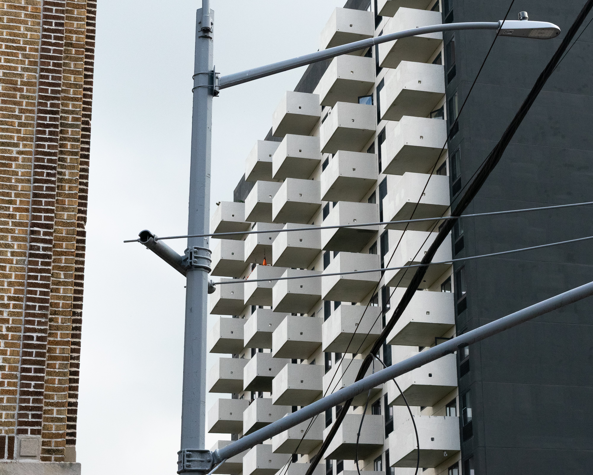 A high-rise apartment tower with rows of cantilevered white concrete balconies stacked in a staggered grid, seen past a gray streetlight pole and crossing overhead power lines, with the curved yellow-brown brick corner of an older building at left.