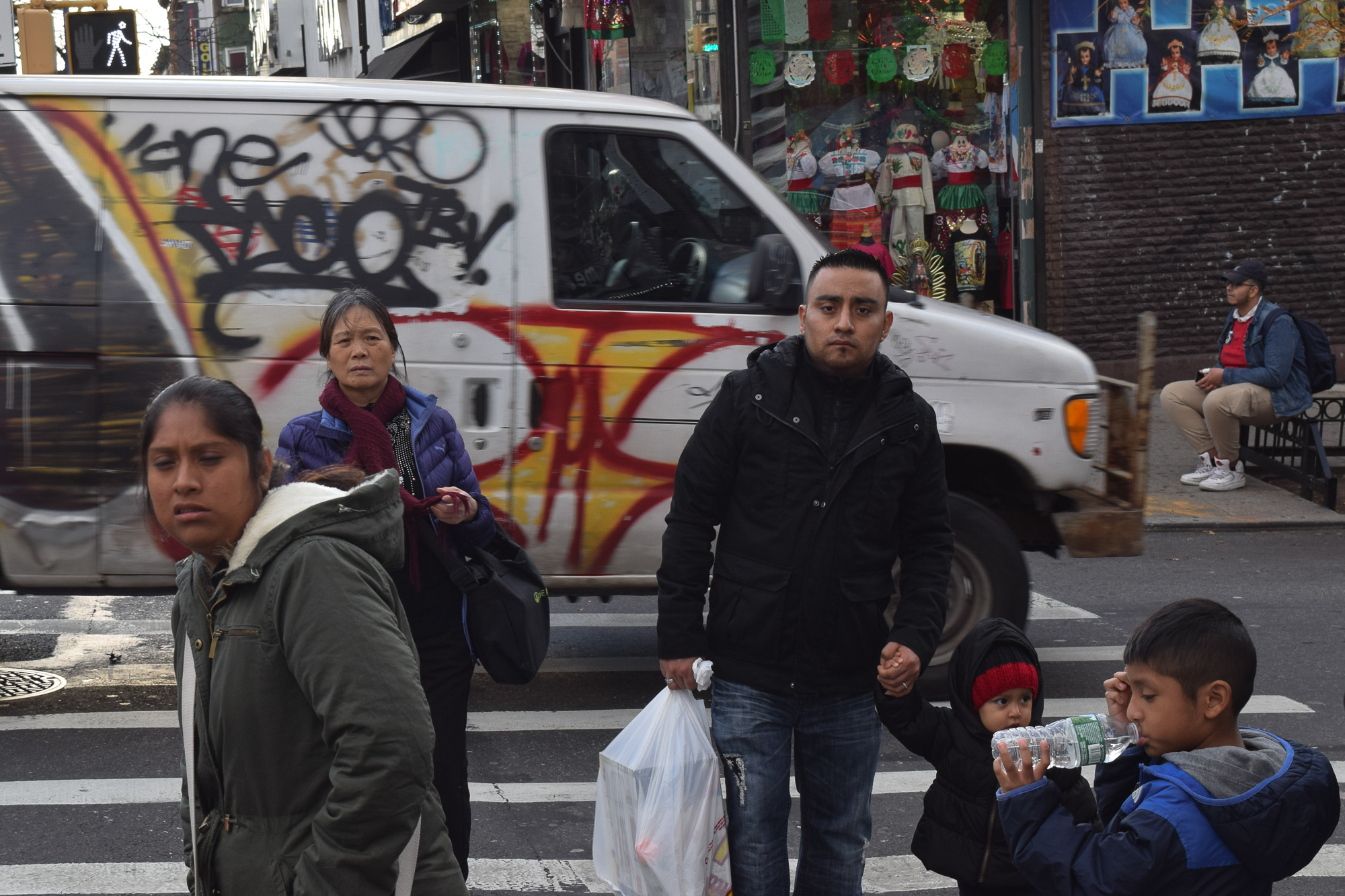 A man in a black hooded coat holds a small child's hand at a crosswalk while a boy in a blue jacket drinks from a water bottle; a woman in an olive parka and another in a purple puffer coat walk nearby as a white van covered in graffiti blurs past behind them, in front of a shop window full of embroidered dresses and religious figurines.