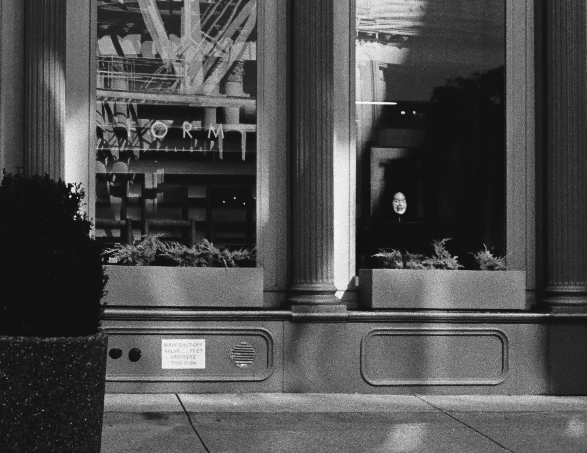 Black and white photograph of a street-level storefront with tall plate glass windows set between fluted stone columns. The left window reflects scaffolding and a partly visible sign reading FORM, while in the dark right window a single pale face appears suspended in the blackness above a planter of ferns. A small placard below reads MAIN SHUT-OFF VALVE, OPPOSITE THIS SIGN.