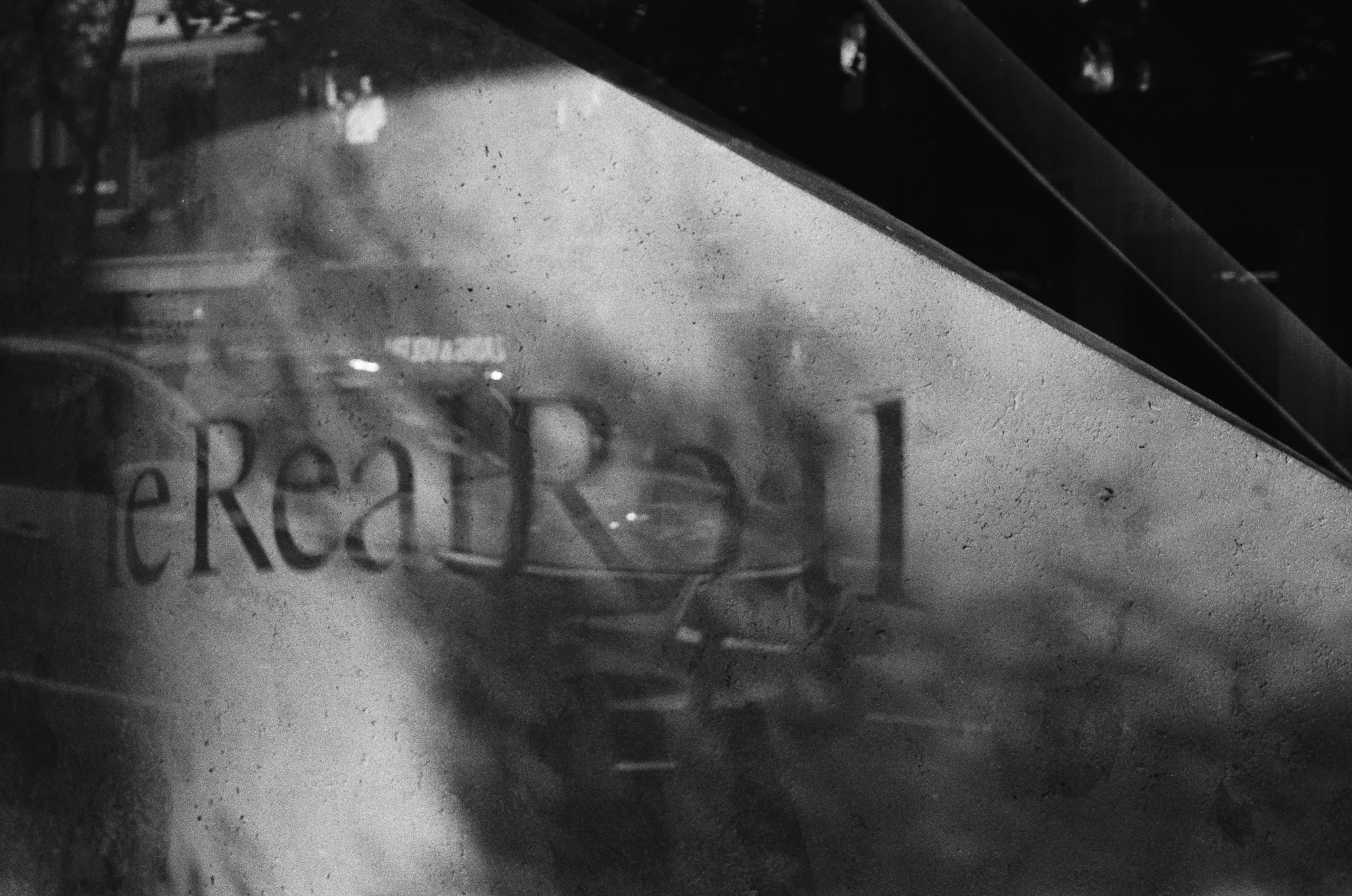 A black and white photograph looking through a hazy, condensation-streaked shop window at serif lettering reading 'heRealReal' on the interior wall, with a dark diagonal band of shadow cutting across the upper right corner and faint reflections of street lamps and passing figures floating across the glass.