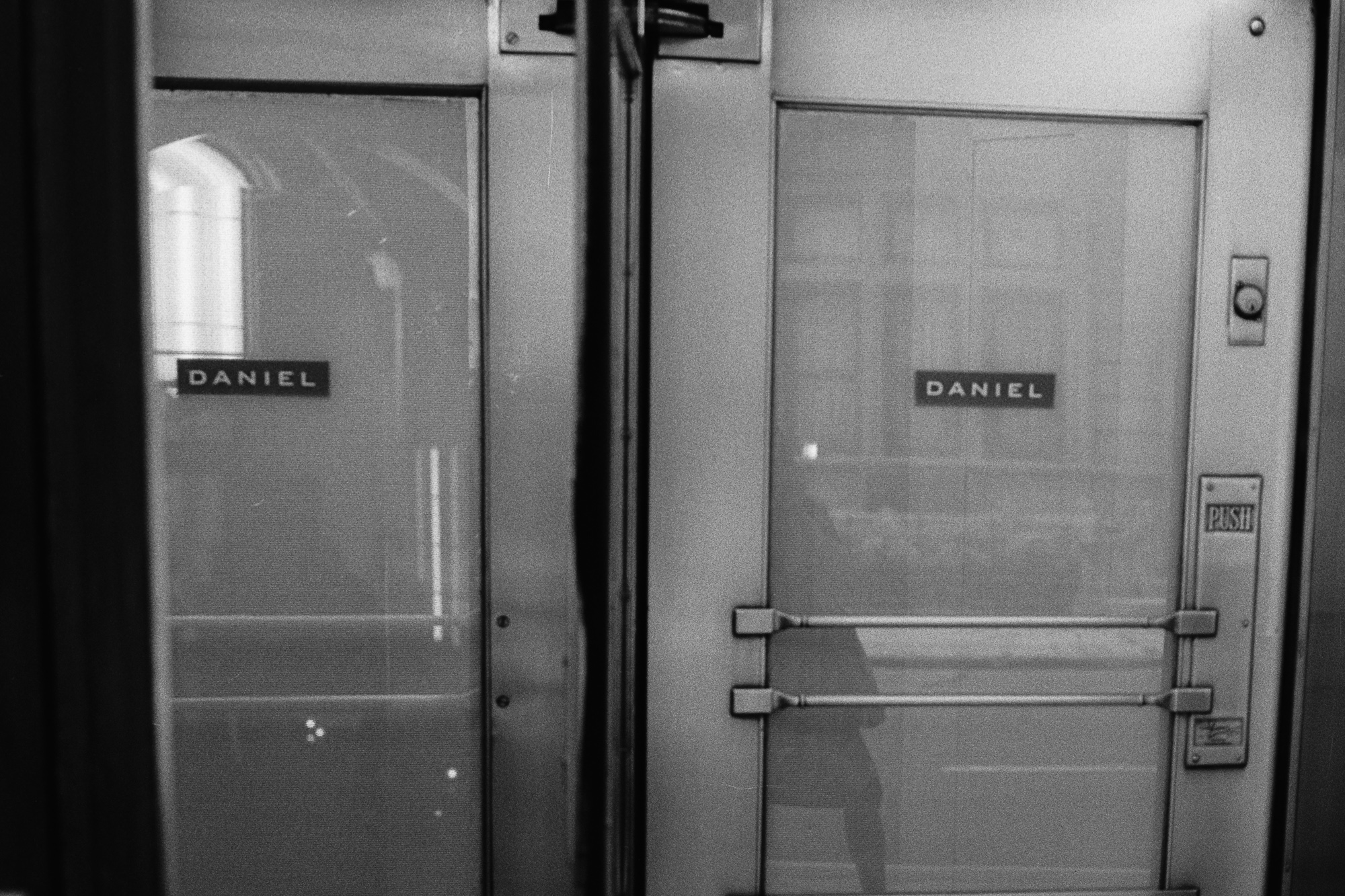 Black and white photograph of two adjacent glass entry doors, each bearing a small dark plaque reading DANIEL in spaced capital letters. The right door has horizontal push bars and a metal plate stamped PUSH, with the arched window and interior of the building reflected across both panes.