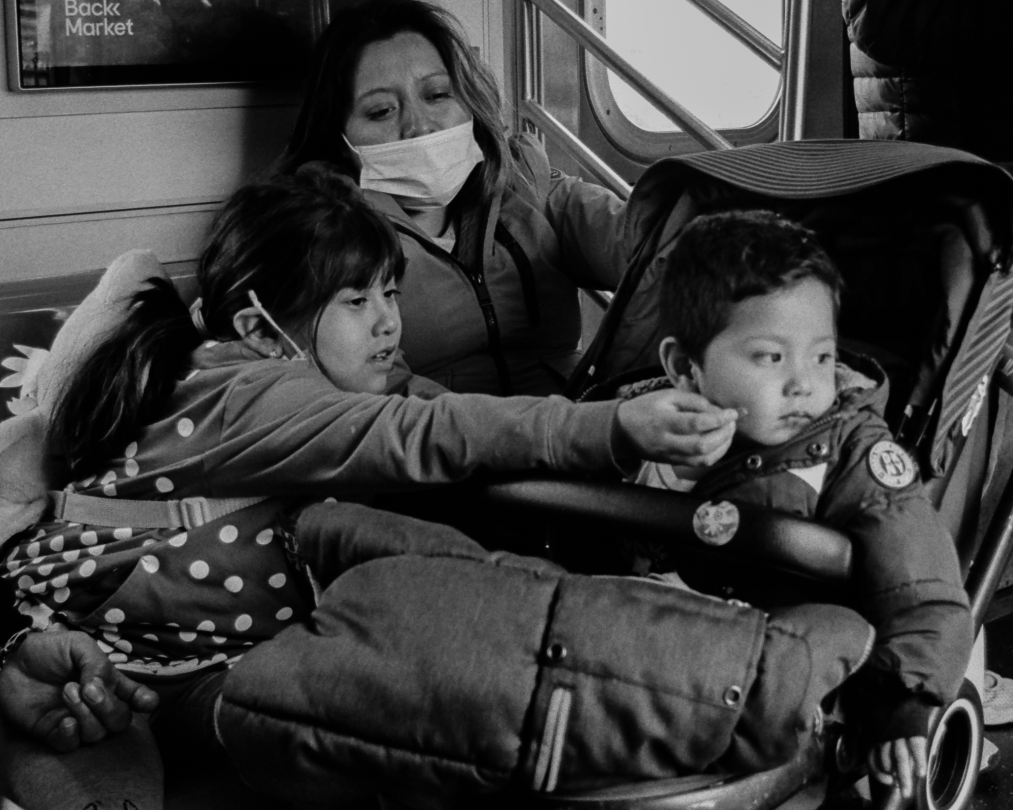 Black and white photo inside a New York subway car: a young girl in a polka-dot dress leans forward with both arms extended to wipe the face of a small boy seated in a stroller, while a woman in a face mask and hooded jacket watches from the seat behind them.