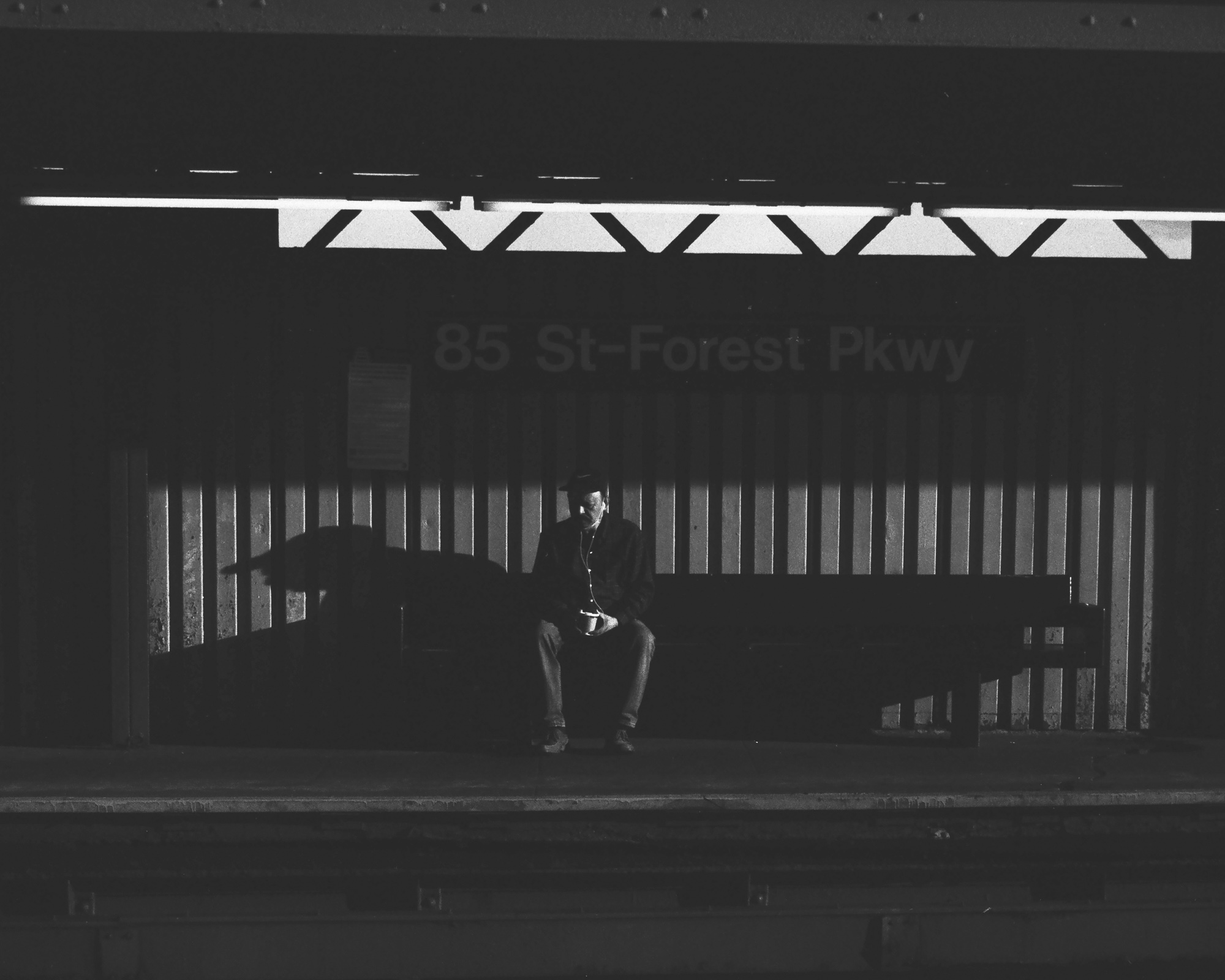 A man in a flat cap and dark jacket sits alone on a bench on a dimly lit elevated subway platform, holding a paper cup in his lap and looking down. A bright fluorescent strip light above throws a zigzag pattern across the ceiling beam, and the station name 85 St-Forest Pkwy is barely legible on the wall behind him.