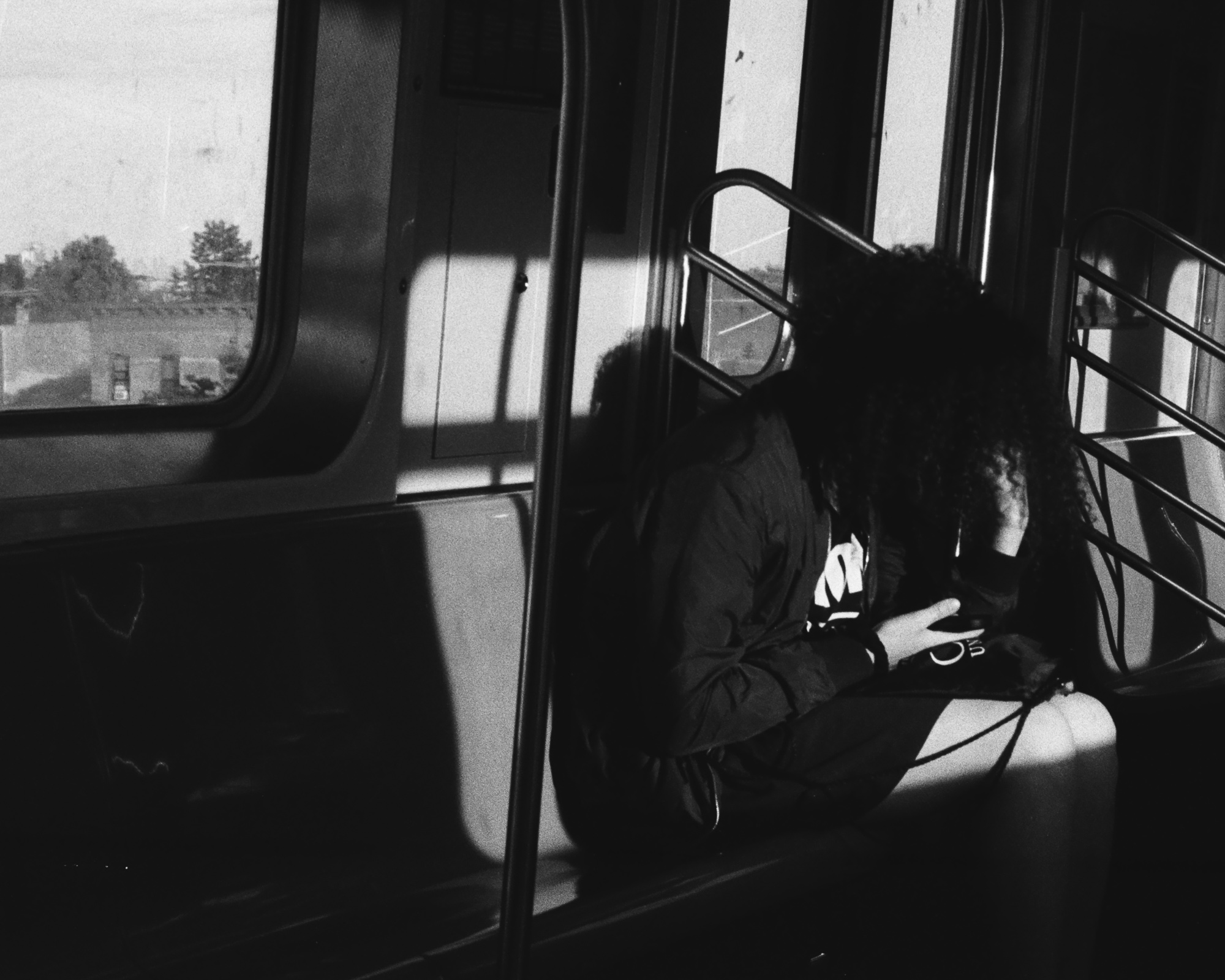 Black and white photo inside a subway car where a person with voluminous curly hair sleeps slumped forward in a dark jacket, head bowed toward their chest, as a hard diagonal band of sunlight cuts across the wall and seat beside them. Through the windows behind, trees and low rooftops pass on an elevated stretch of track.
