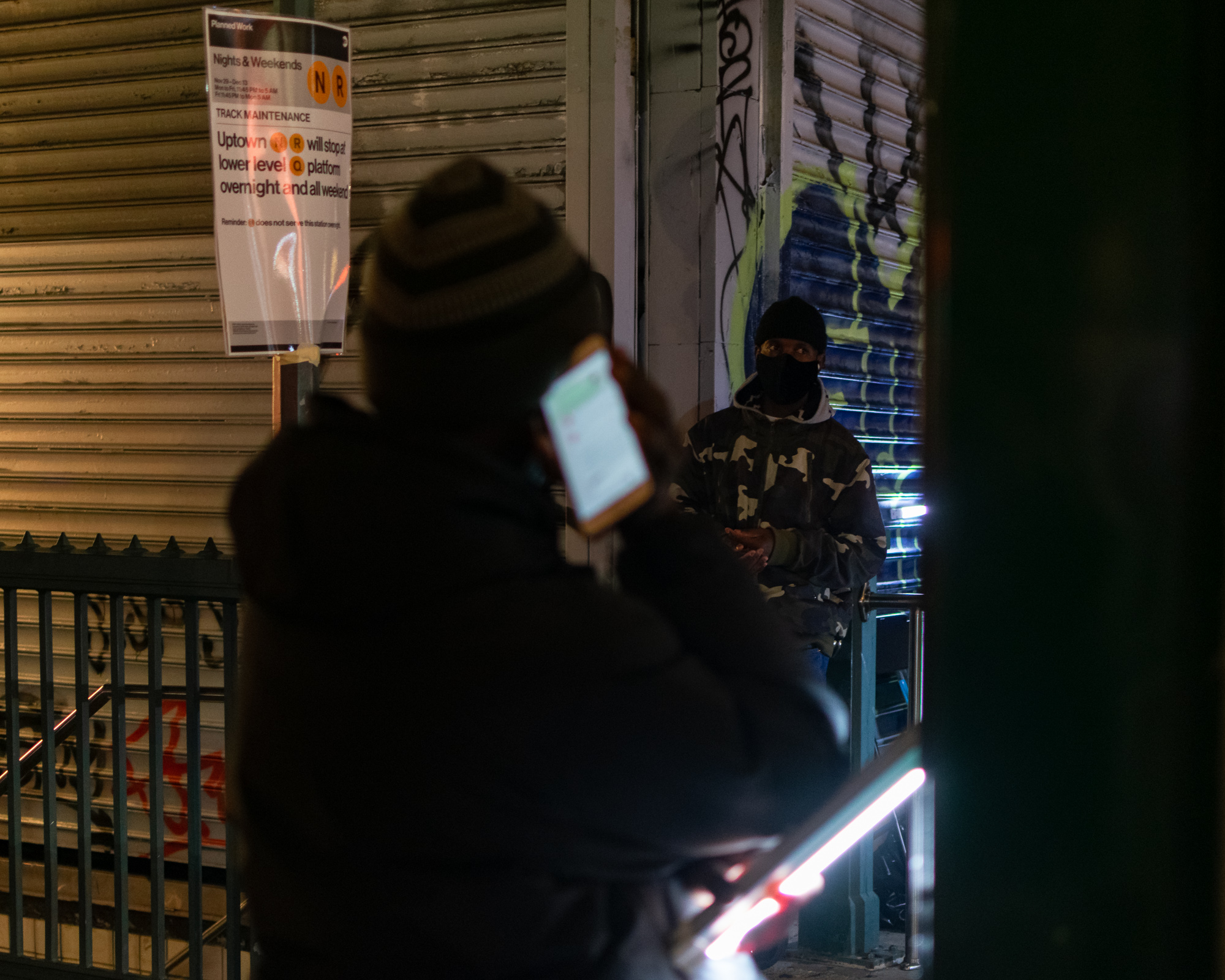 At night outside a subway entrance, a person in a knit cap seen from behind holds a glowing phone to their ear, blurred in the foreground. Behind them a masked man in a camouflage jacket leans against a graffiti-covered roll-down gate beside an MTA Planned Work poster announcing N and R track maintenance.