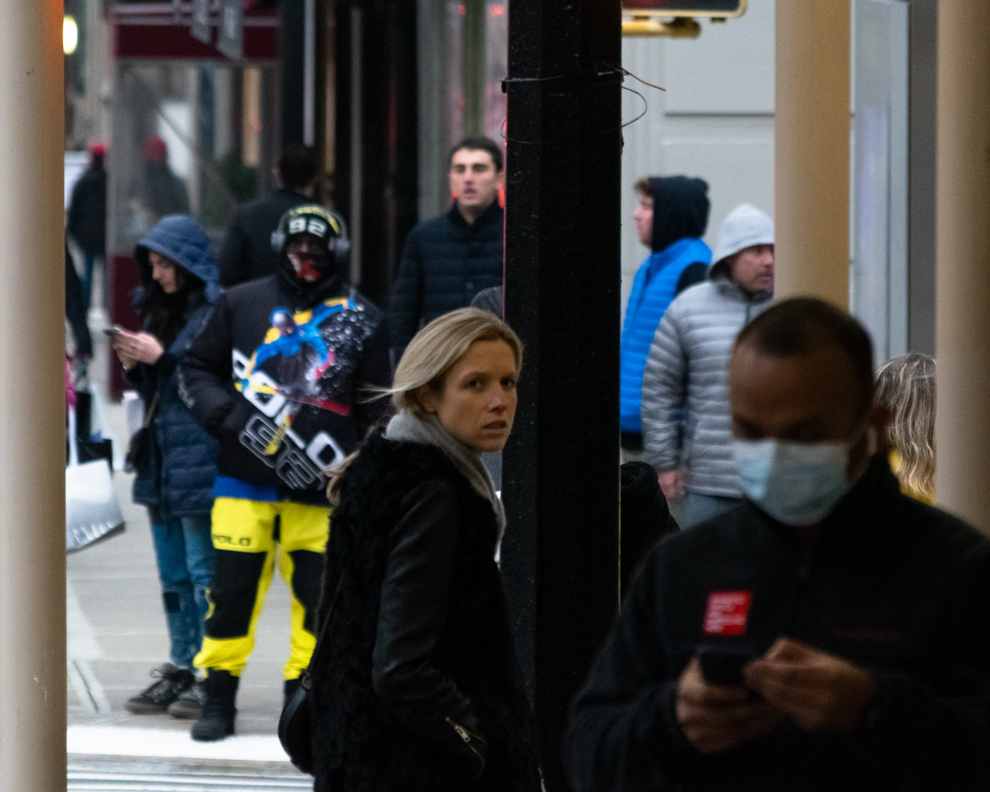 On a crowded Manhattan sidewalk, a woman in a black fur-collared coat and grey scarf turns her head to look directly at the camera from behind a black lamppost, while a man in a surgical mask checks his phone in the right foreground and pedestrians in puffer jackets, including one in bright yellow Polo snow gear, fill the background.
